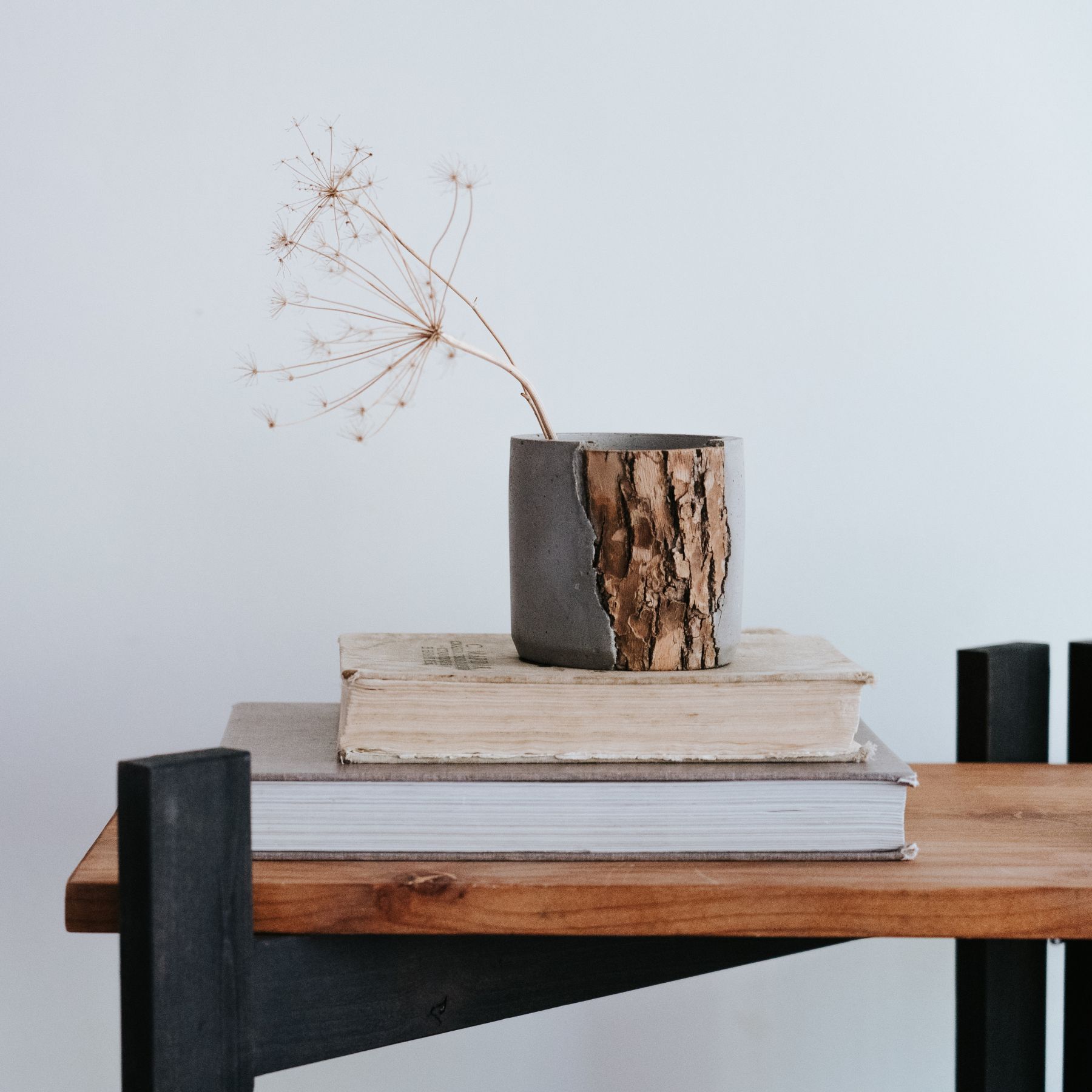 Minimalist vase with bark texture holding a dried flower, placed on two stacked books on a wooden table.