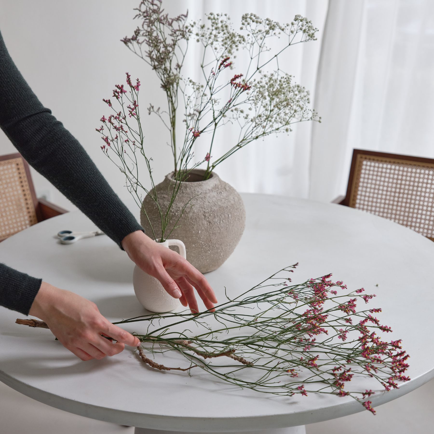 Hands arranging delicate pink and white flowers on a white round table with two vases and cane-back chairs in the background.
