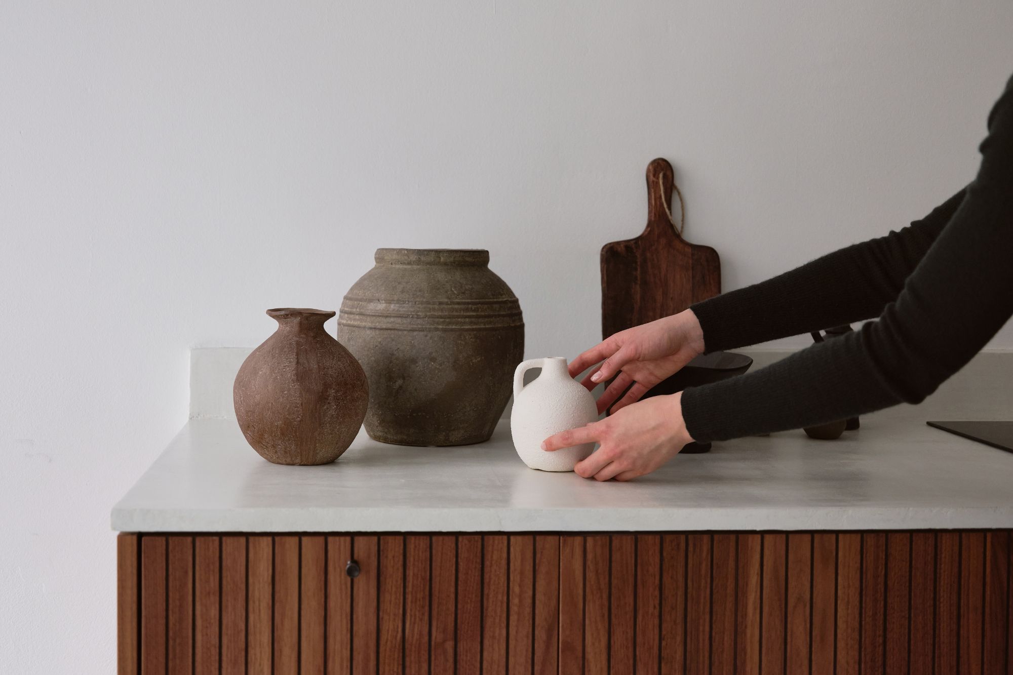 Hands placing a small white ceramic jug on a countertop with two larger rustic clay pots and a wooden cutting board in the background.