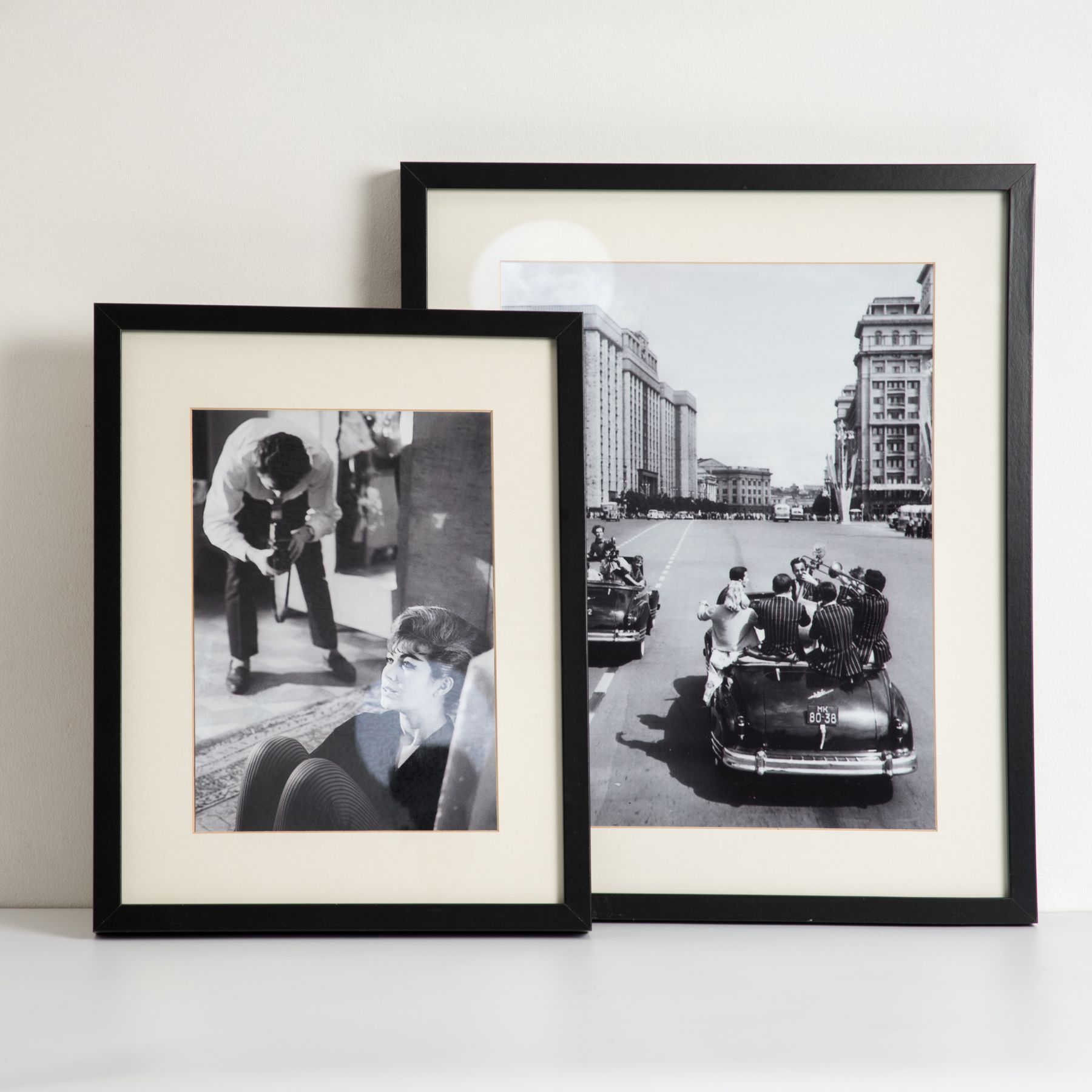 Two black-framed black and white photos, one showing a person photographing a seated woman indoors, the other showing a group of people playing instruments on a car in a city street.