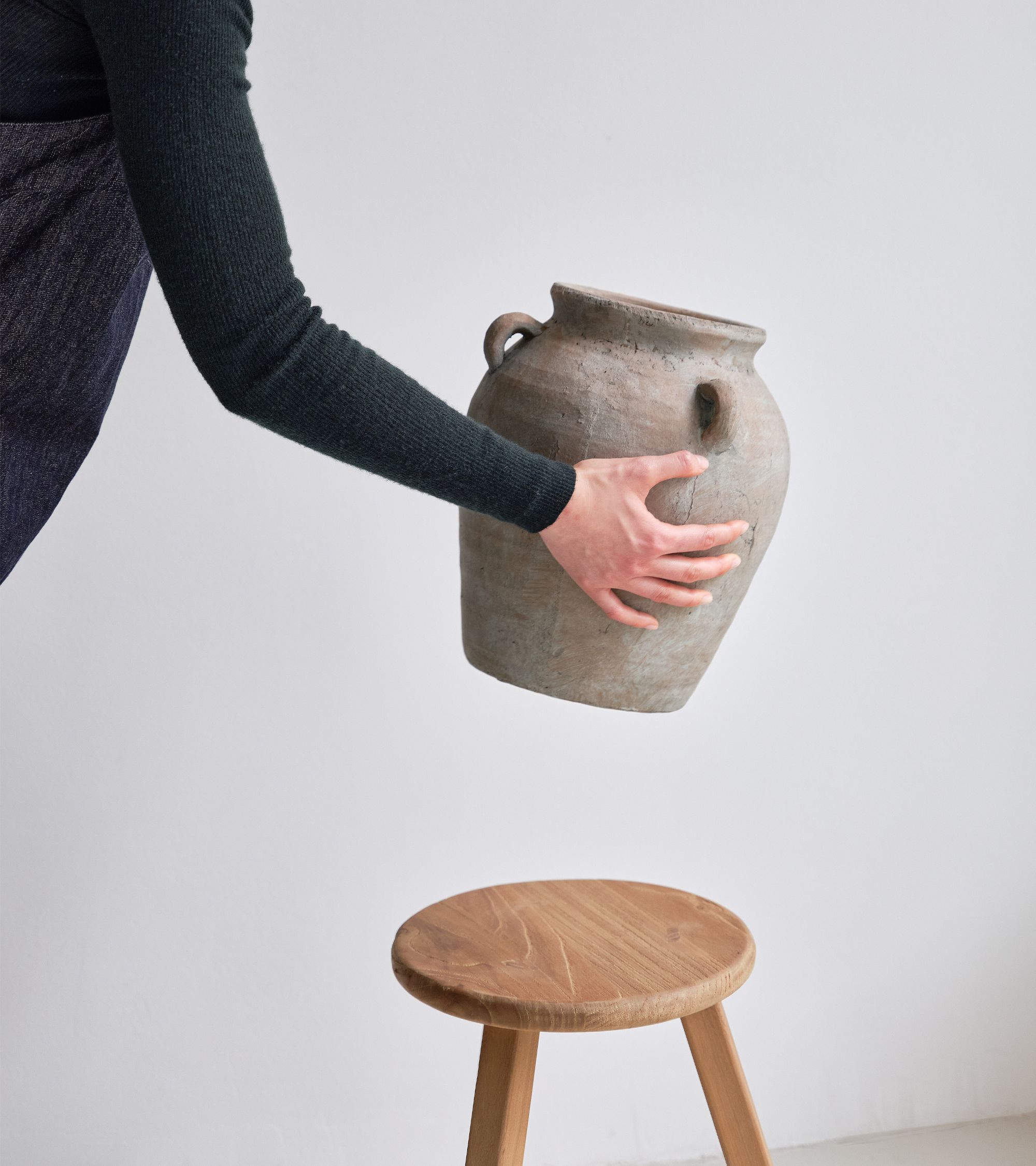 Person wearing a dark sweater holding a clay pot above a wooden stool against a plain white wall.
