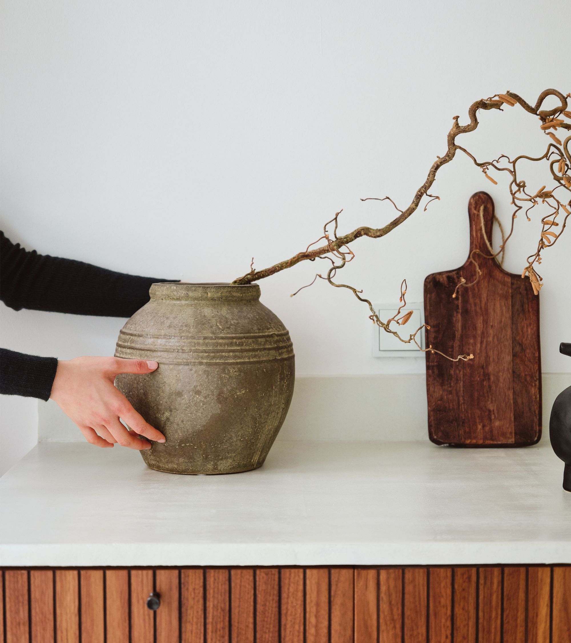 Hands placing a large rustic ceramic vase with a dry twisted branch on a white kitchen countertop, with a wooden cutting board in the background.
