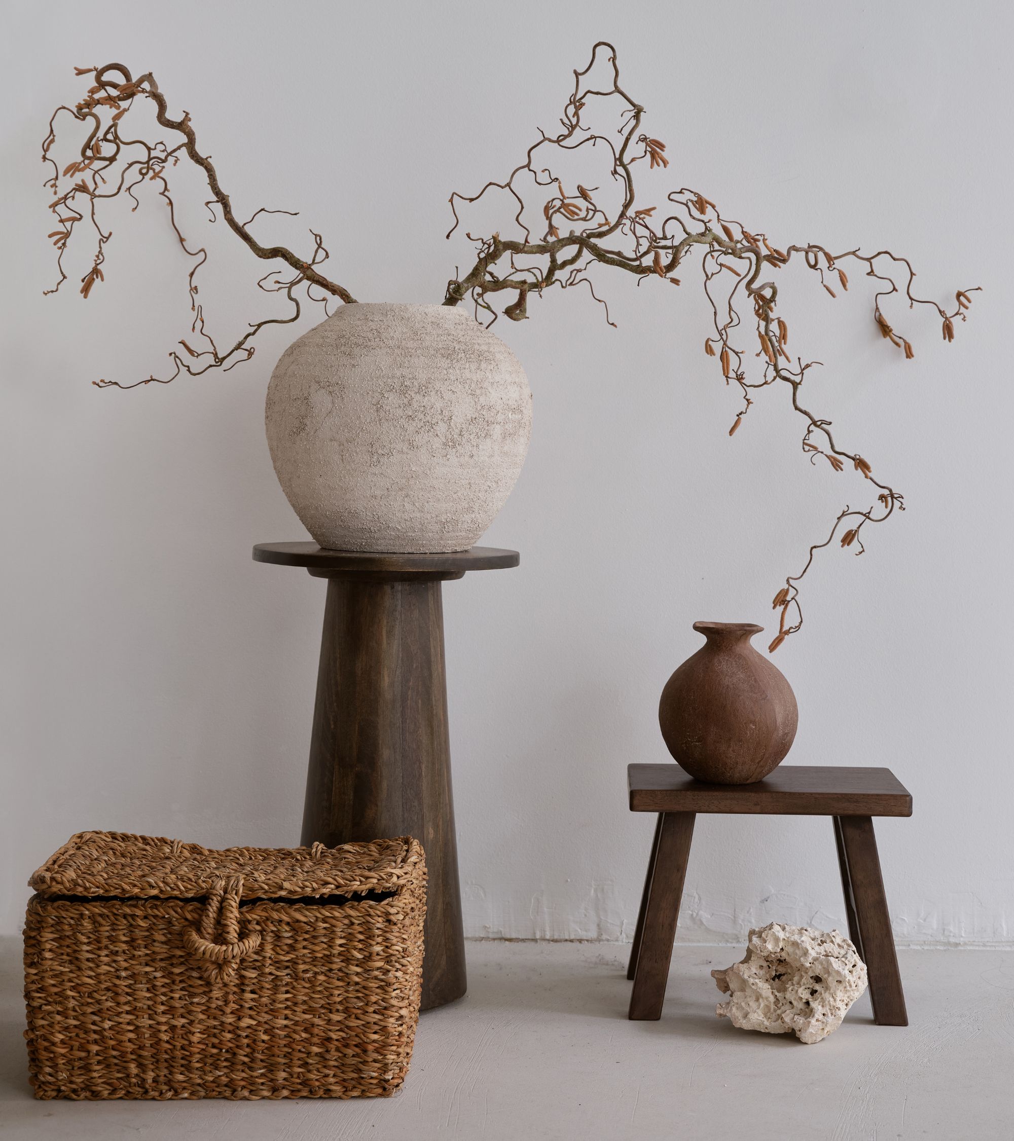 Two brown vases with twisted bare branches placed on dark wooden stands next to a woven basket and a white rock on a white floor in front of a light wall.