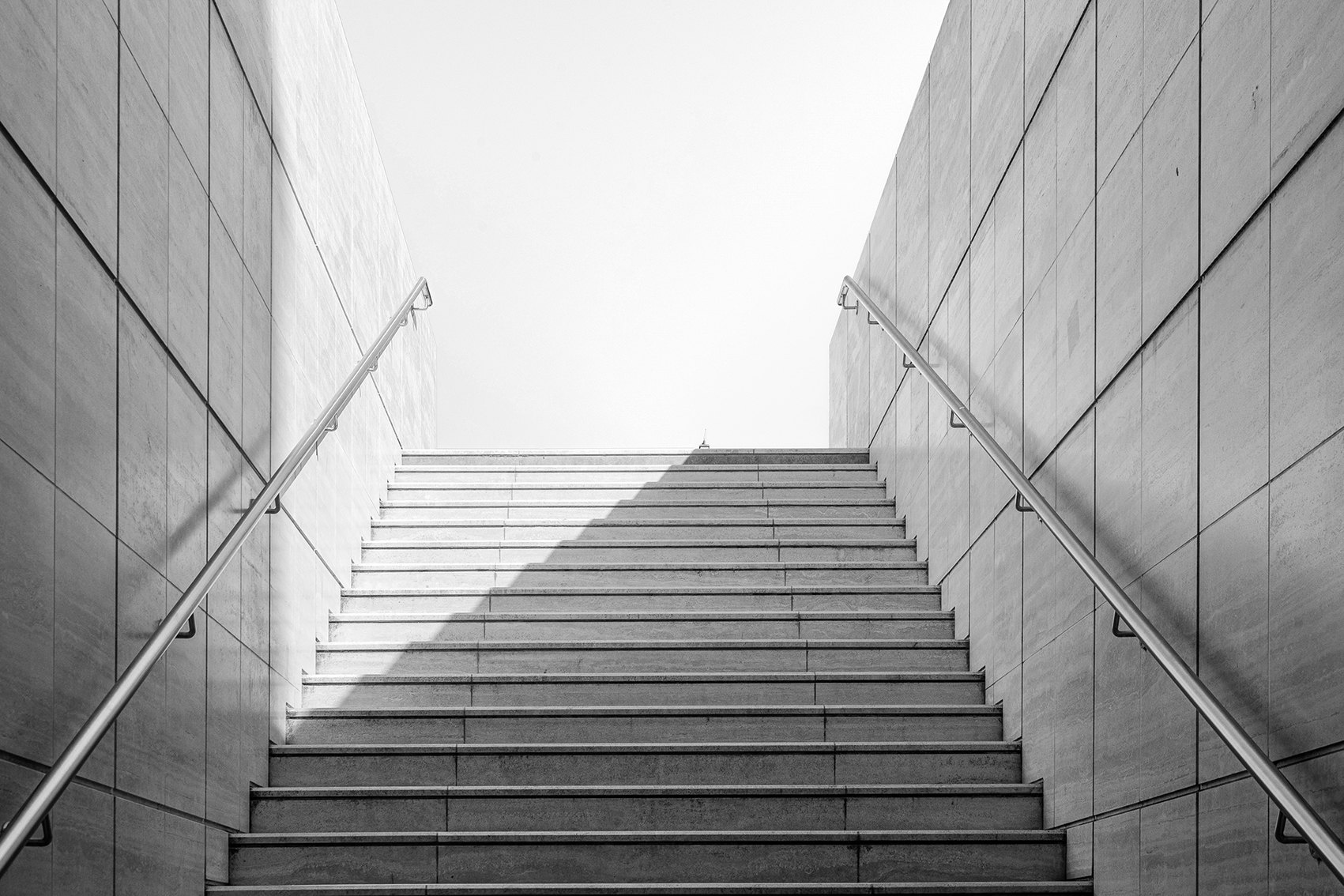 Image d'un escalier en pierre avec rampes métalliques de chaque côté, entre deux murs carrelés, éclairé par la lumière naturelle. 