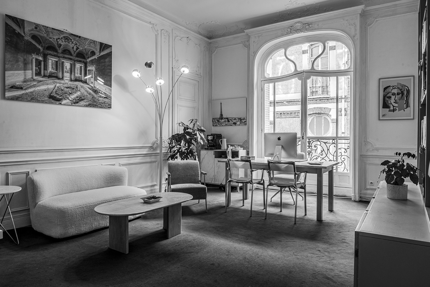 Photo illustrant un bureau élégant avec canapé, fauteuil, table basse, bureau en bois avec ordinateur iMac, grandes fenêtres avec balcon et décoration murale.