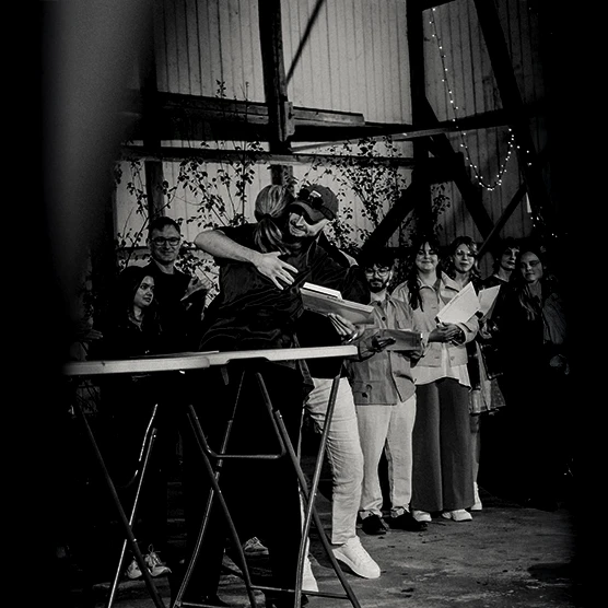 A black and white photo of two people hugging near a table, surrounded by a group of people standing and watching in what appears to be an indoor event space decorated with string lights.