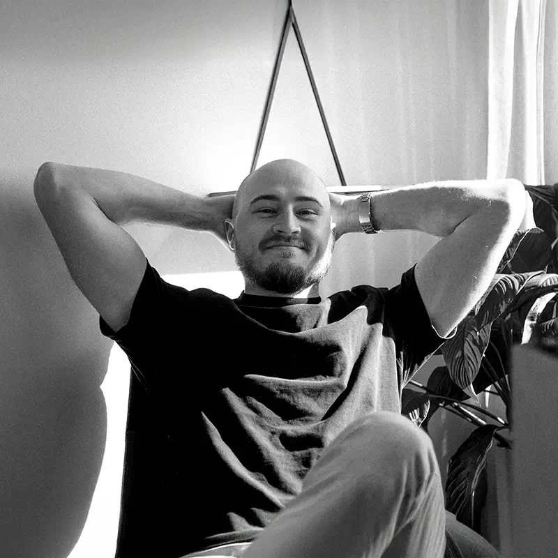 Johan with a shaved head and beard sits relaxed in a chair, arms resting behind his head, smiling. Sunlight streams through a window, and a leafy plant is visible beside him. The photo is in black and white.