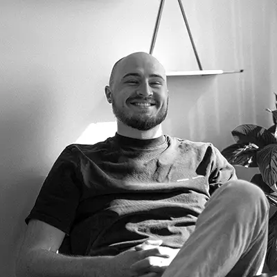 Smiling bald man with a beard sitting comfortably in a chair in a well-lit room with a plant nearby.