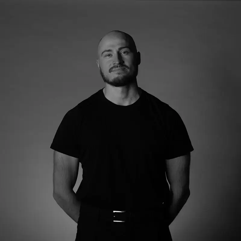 Johan with a beard, wearing a black t-shirt, stands with his hands behind his back against a plain, dark background. The image is in black and white.