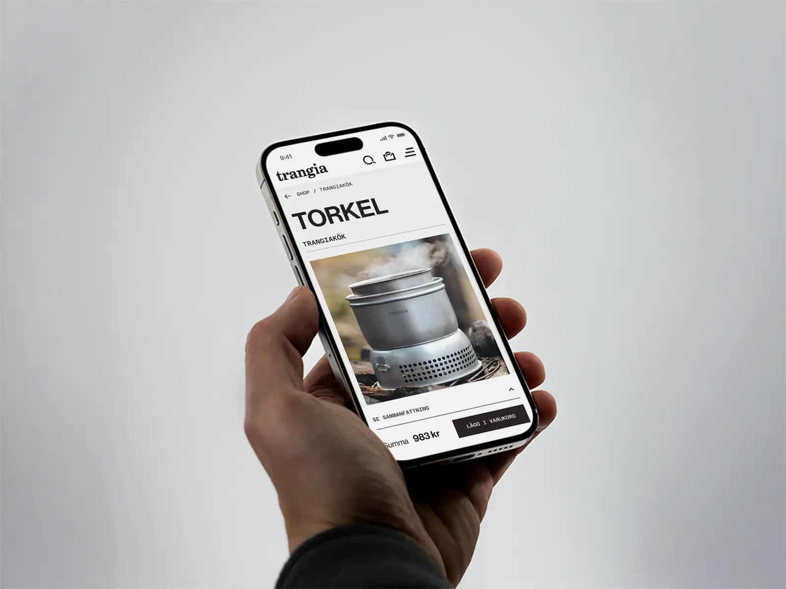 A hand holds a smartphone displaying a product page for a portable camping stove called Torkel on the Trangia website. The stove is shown emitting steam or smoke.