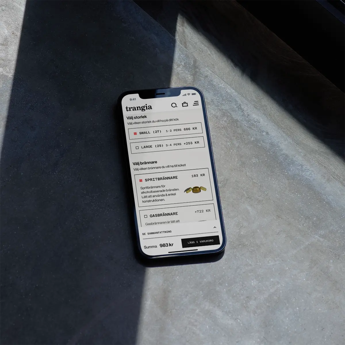 A smartphone lying on a gray surface displays a shopping website in Swedish, showing options for Trangia stove kits with prices and product details under natural light.