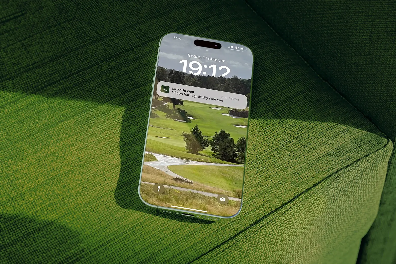 A smartphone with a golf course wallpaper and a notification from Links'Up Golf rests on a textured green fabric surface, possibly a couch or chair. 