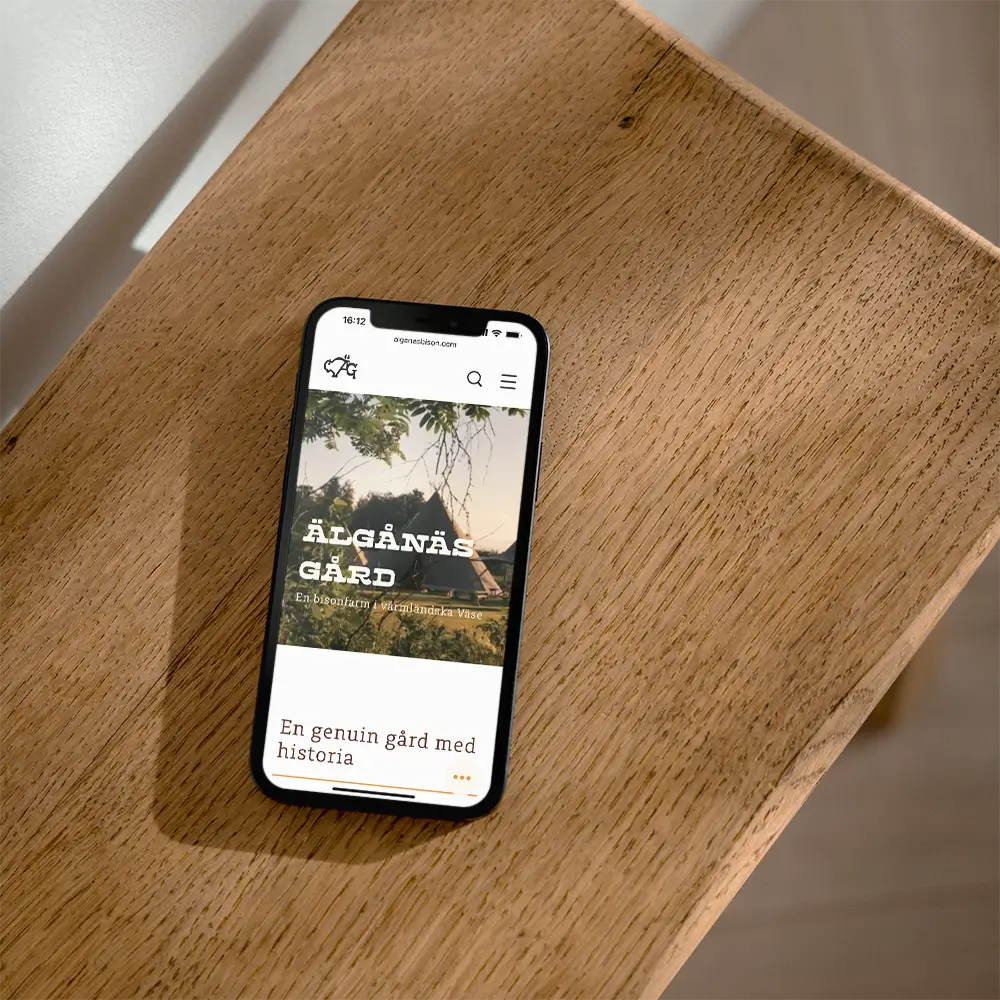 A smartphone displaying a website featuring Södra Älgånäs Gård rests on a wooden surface, with part of a chair and floor visible in the background.