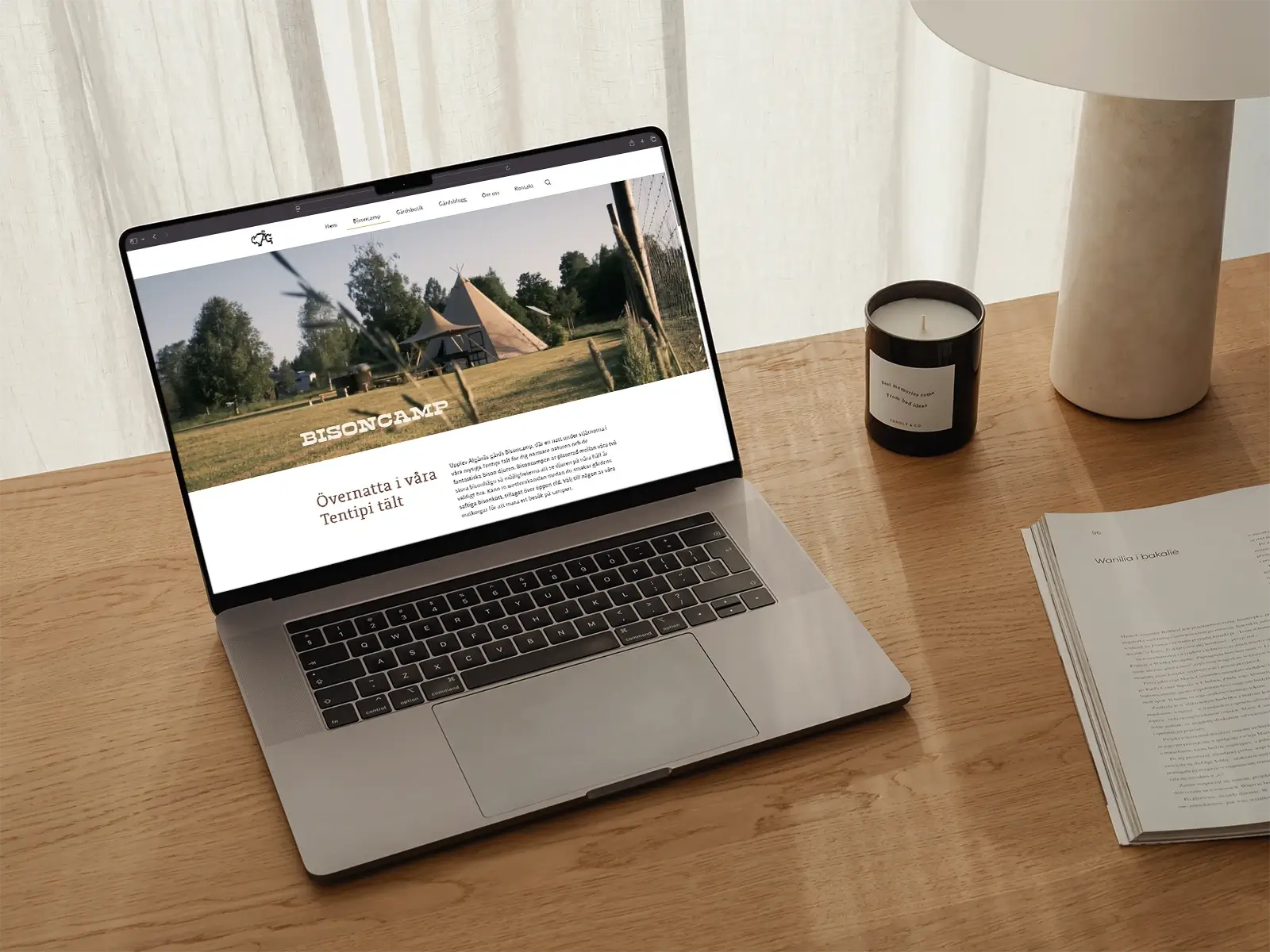 A laptop on a wooden desk displays a website for BisonCamp featuring tipi tents and trees. Nearby are a candle, a stack of papers, and a white lamp. Light filters through a curtain in the background.