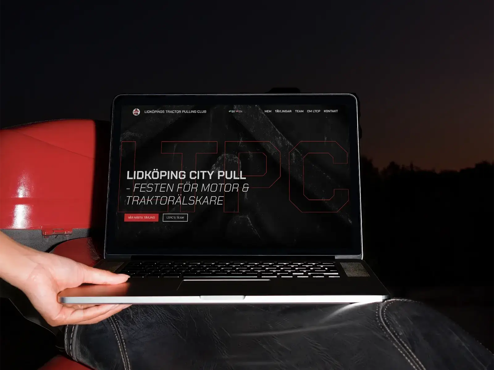 A person holds a laptop outside at dusk, displaying a website for Lidköping City Pull, an event for tractor enthusiasts. The screen text is in Swedish, and the laptop rests on a red surface, likely a tractor.