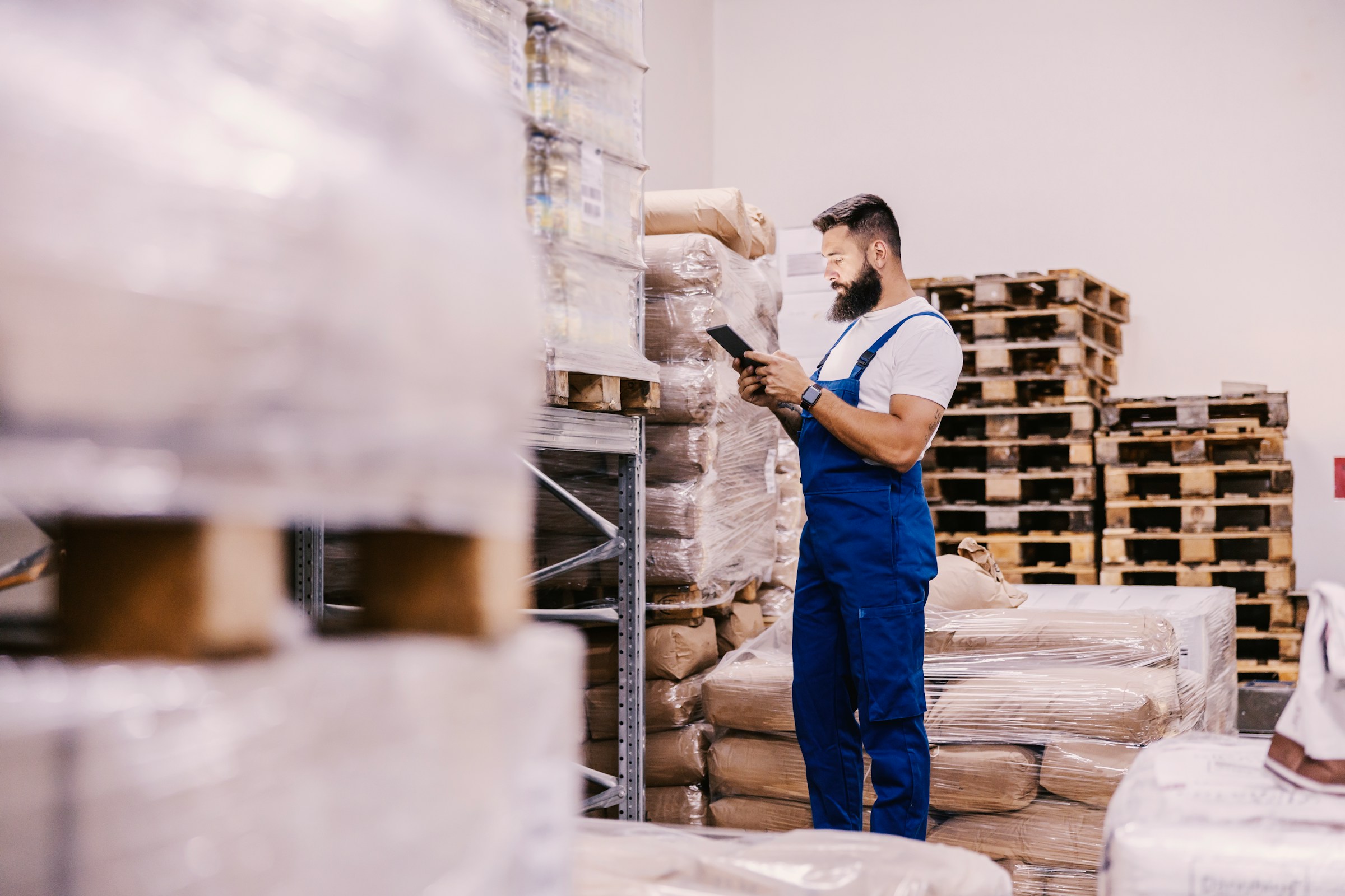 Man in blue overalls using a tablet while standing in a warehouse surrounded by stacked pallets and wrapped packages.