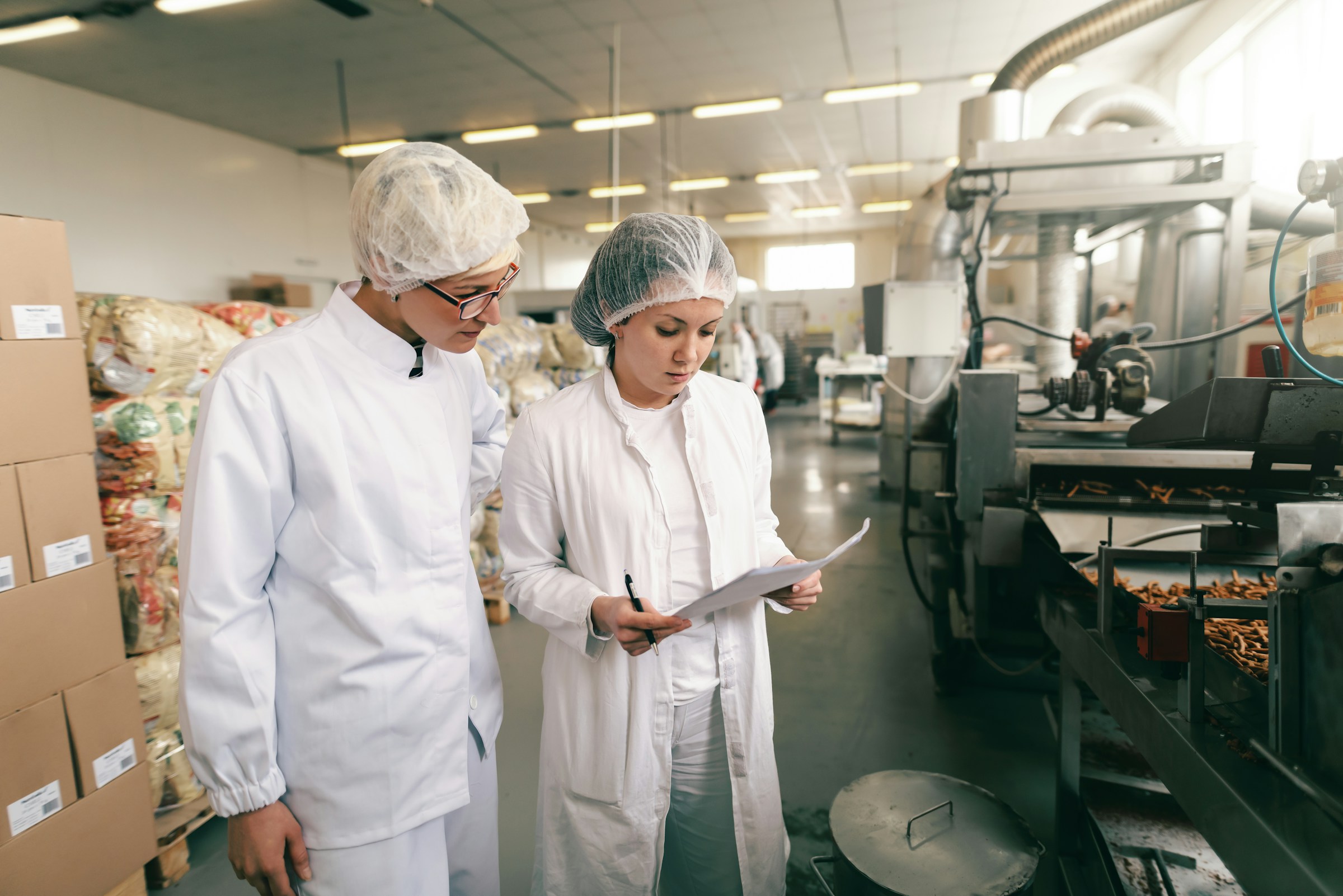 Two food factory workers wearing white coats and hair nets reviewing documents by a production line filled with pretzels.