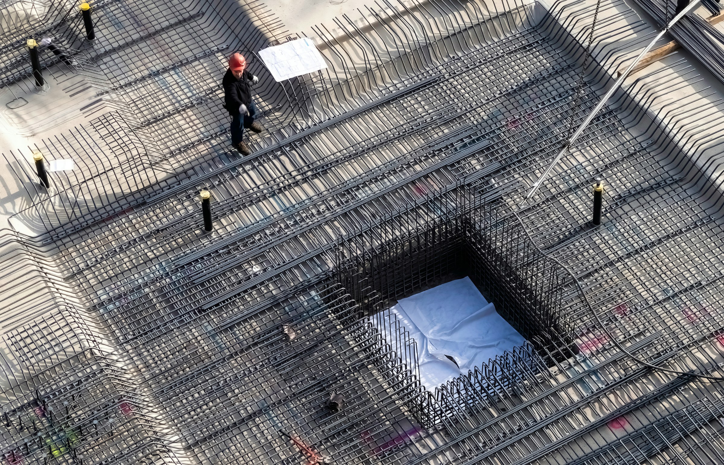 Construction worker wearing a red hard hat standing on steel rebar grid framework at a building foundation site with concrete base.