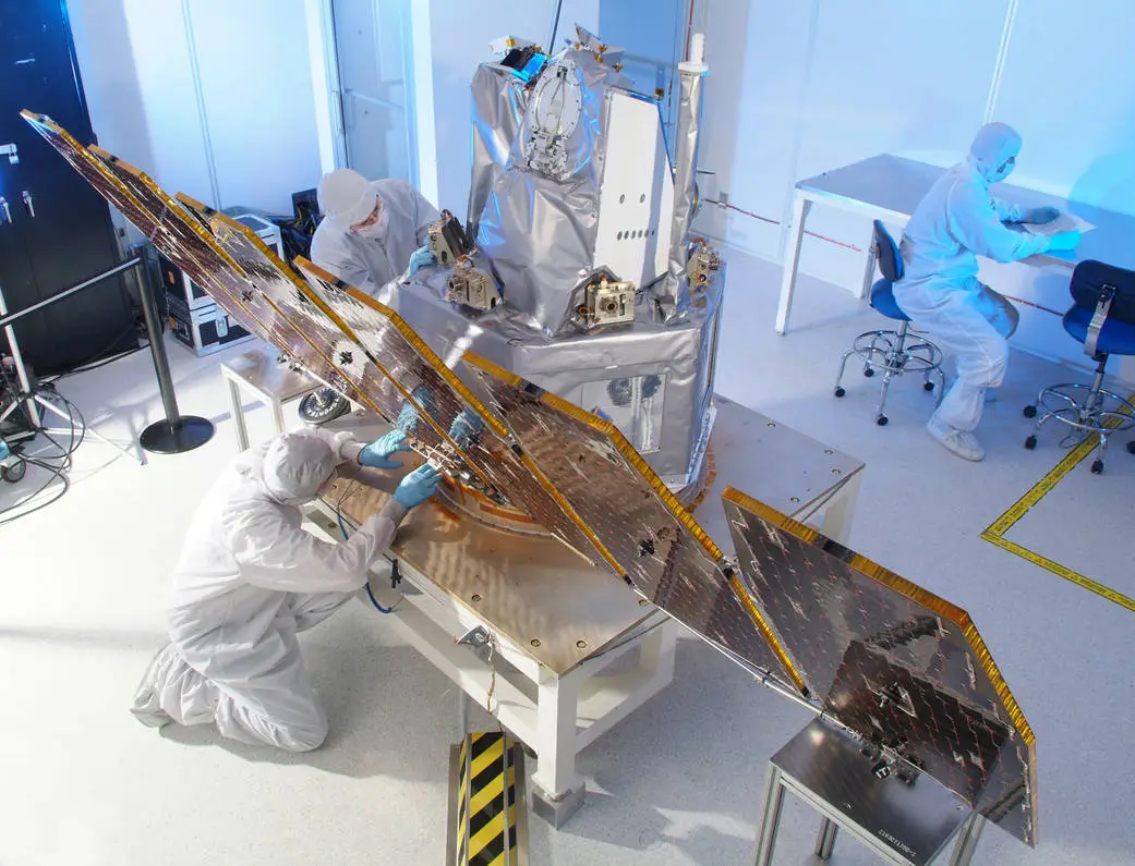 Technicians in cleanroom suits assembling and inspecting a spacecraft with large solar panels.