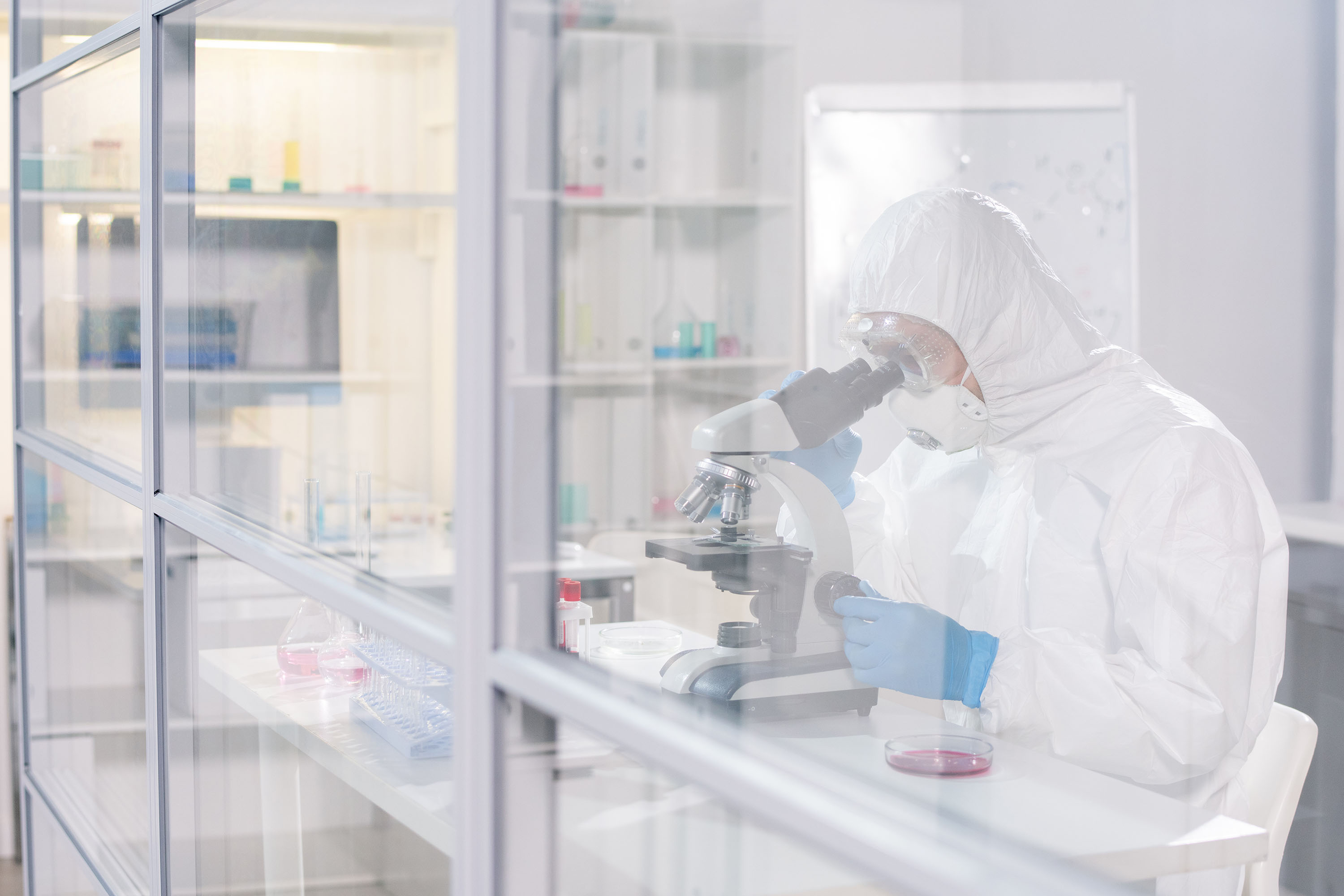 Scientist in full protective suit and gloves looking into a microscope in a laboratory.