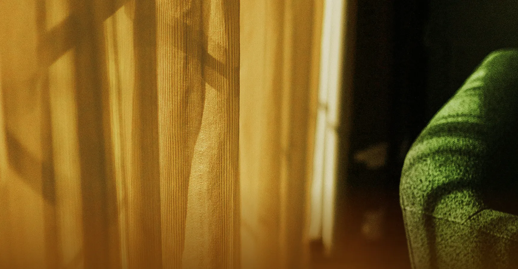 Sunlight casting shadows through a sheer yellow curtain next to the arm of a green textured sofa.