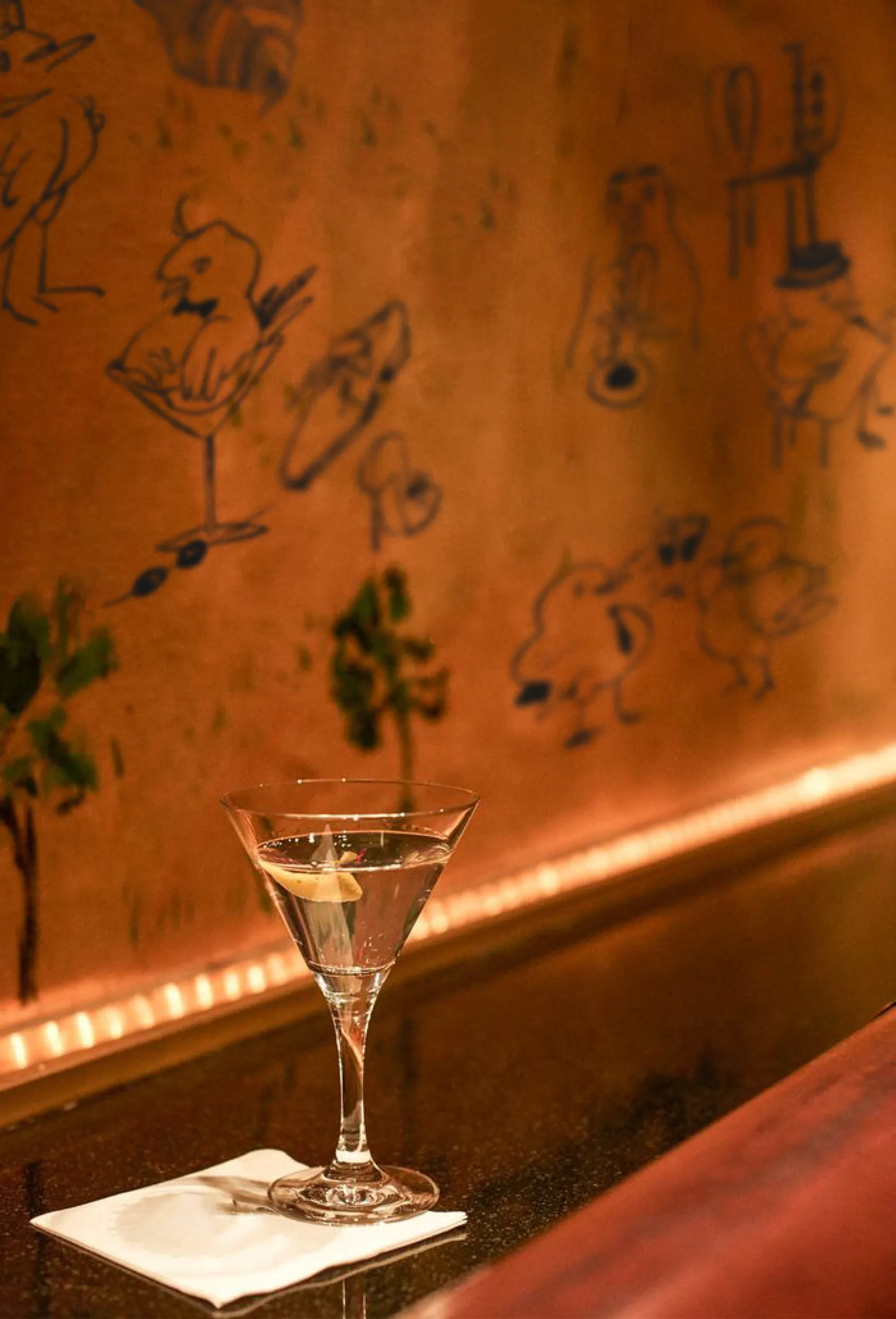 A clear gin martini with olive on a white napkin on a bar counter with bird-themed wall art.
