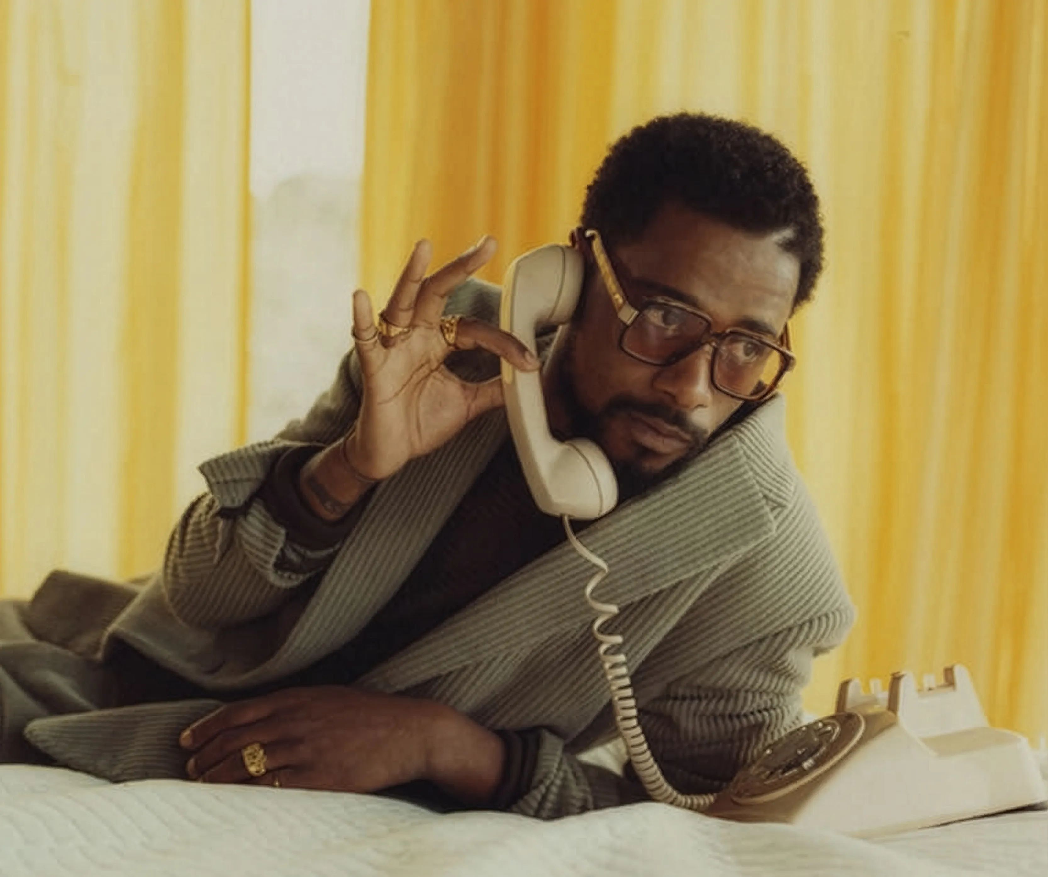 Man with glasses lies on bed holding a rotary phone receiver to his ear near yellow curtains.