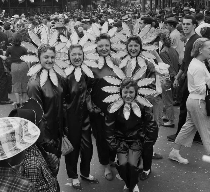 Five women in dark costumes with large flower petal headpieces standing in a crowded street during Mardi Gras 1955.
