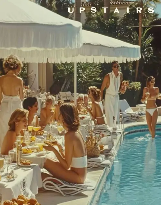 Group of women and a waiter enjoying drinks and food by a pool under white umbrellas.