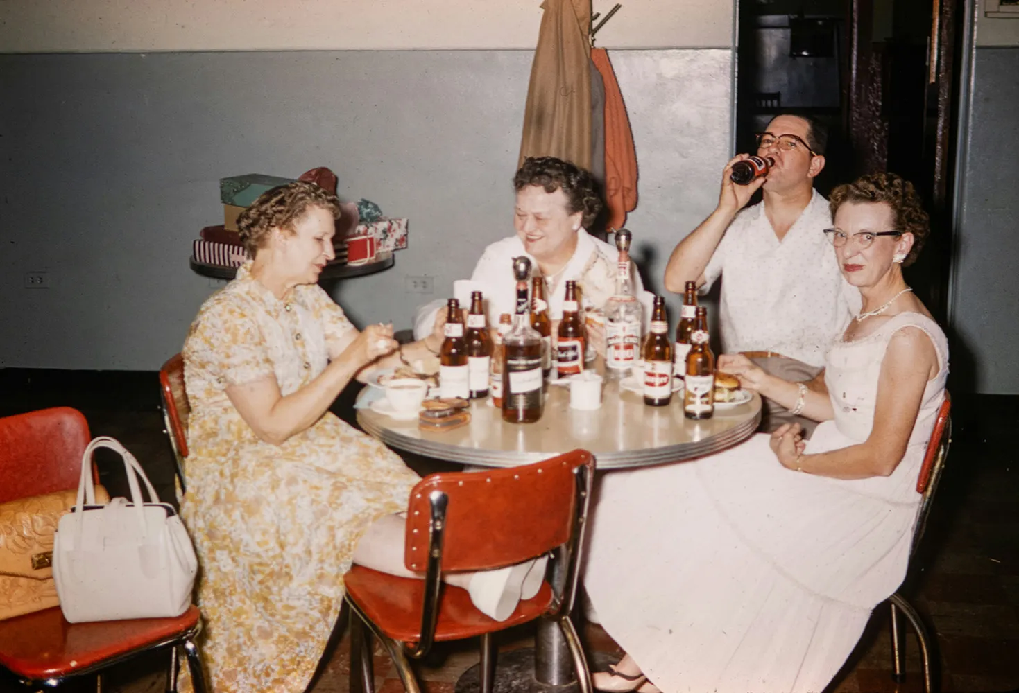 Four adults in vintage clothes sitting at a table with bottles and plates, enjoying a social gathering.