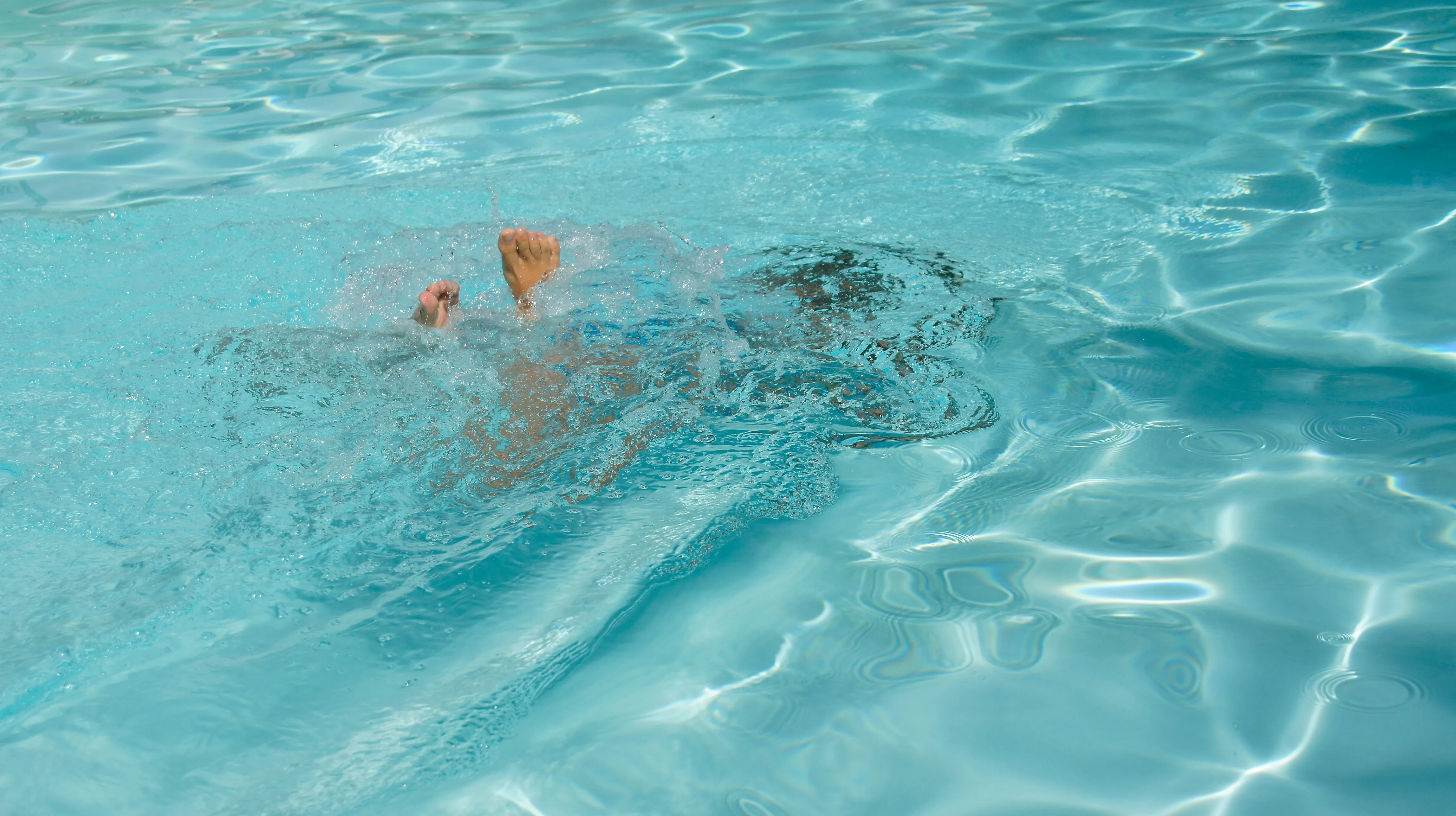 Person submerged underwater in a pool with feet and partial head visible amid splashing water.
