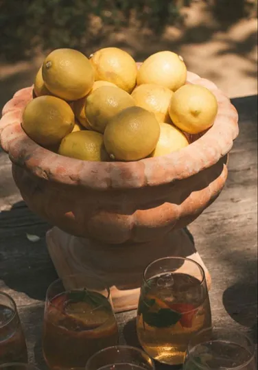 Terracotta bowl full of lemons on a wooden table with glasses of iced drinks nearby.