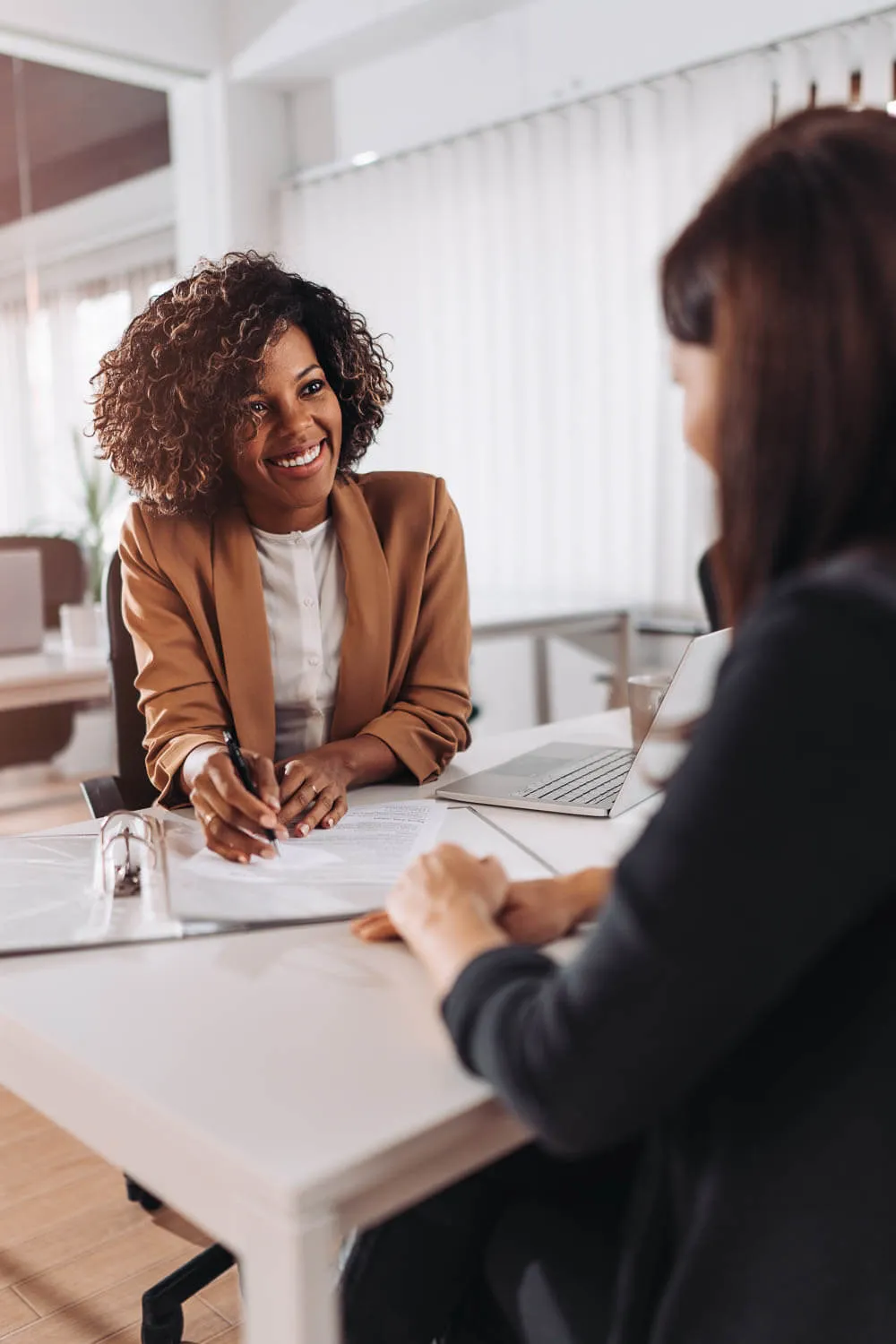 Two women sit across from each other at a desk in an office, one smiling and holding a pen while the other reviews documents.