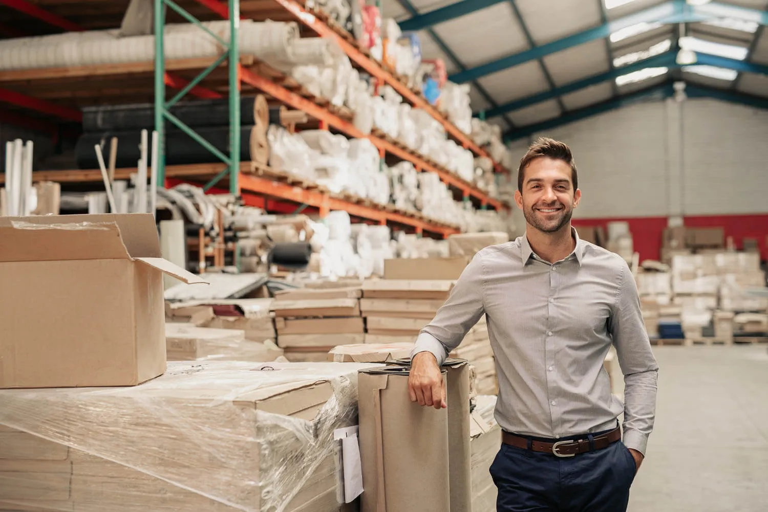 A man in a button-up shirt stands and smiles in a warehouse filled with stacked boxes and shelves.