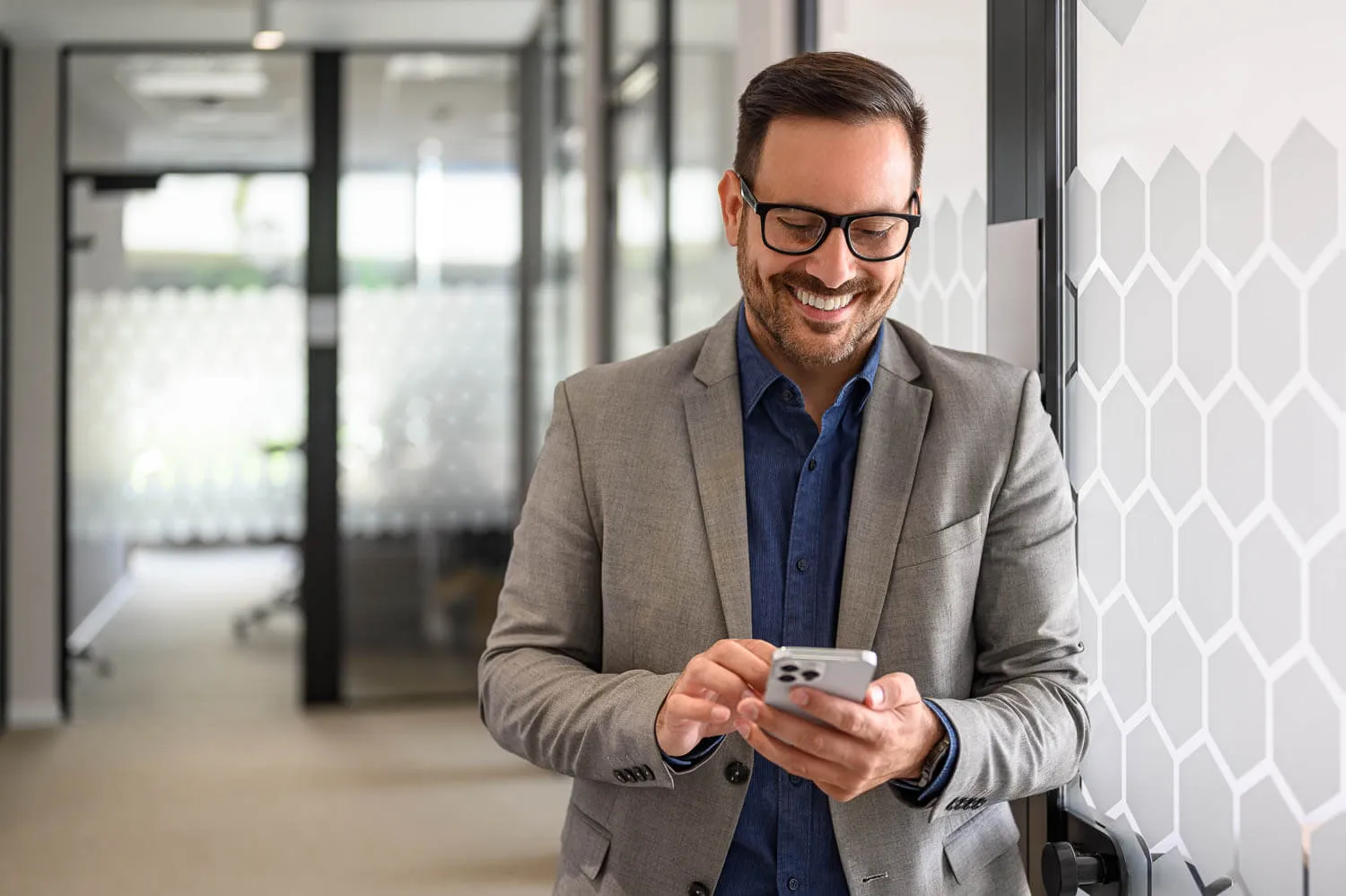 A businessman in a gray suit and glasses stands indoors, smiling while using a smartphone to review his finances.