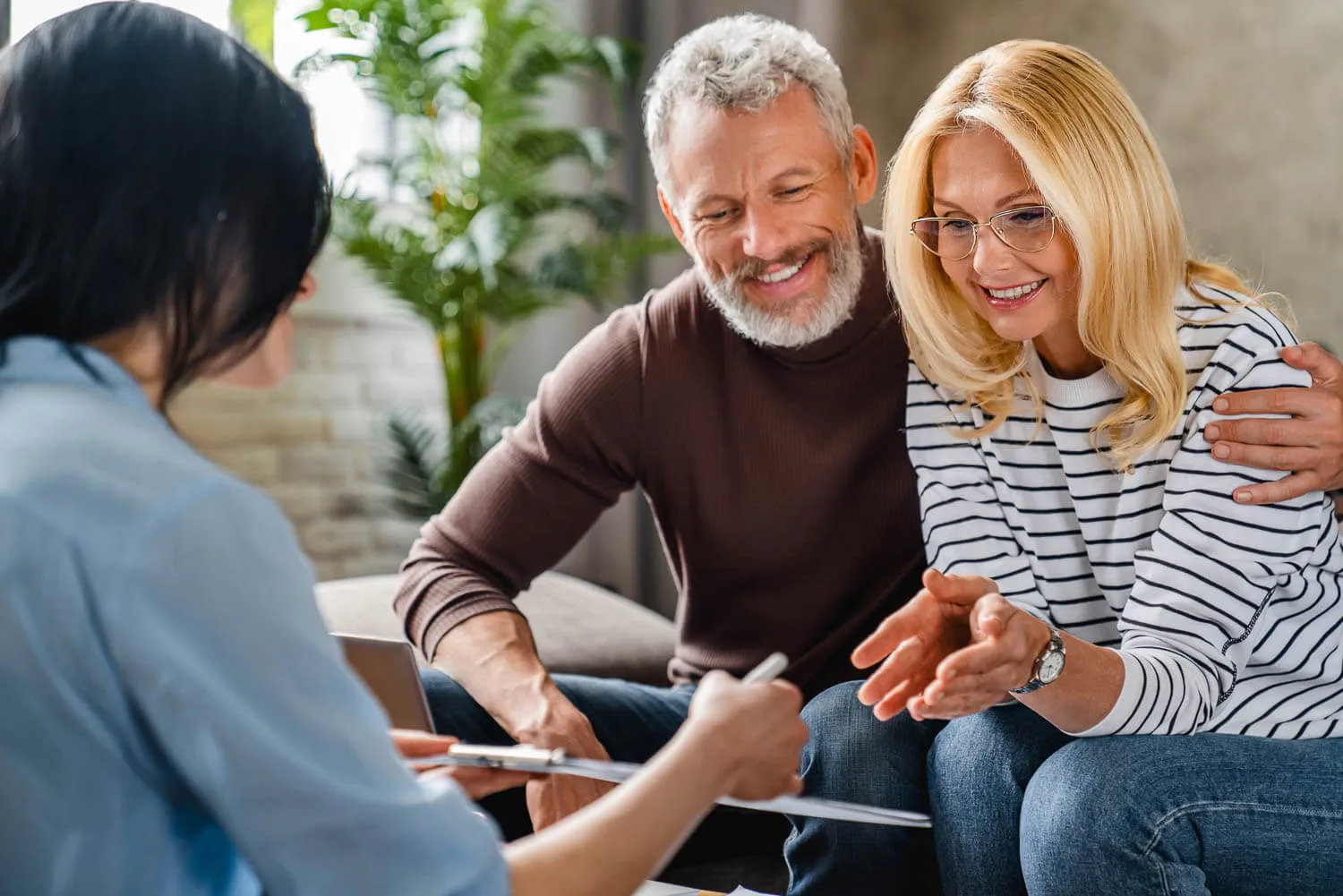 A smiling middle-aged couple sits on a couch, speaking with a professional woman holding their tax report and pen during a consultation.