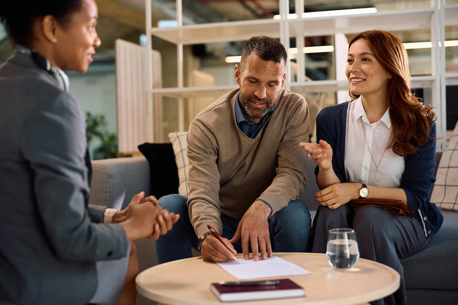 Three middle aged people sit around a coffee table; a man signs a document for the preparer of their annual taxes.