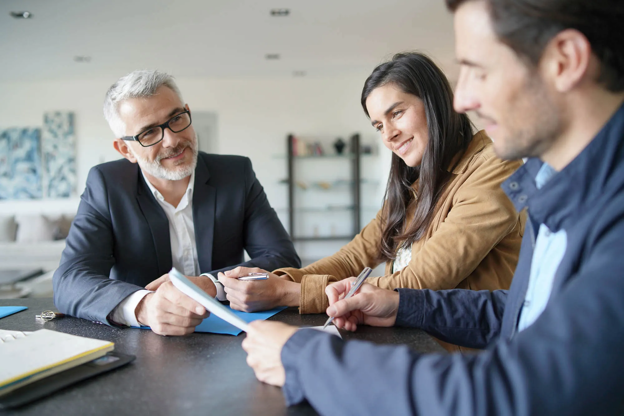 Three people sit at a table discussing tax planning; an accountant in a suit faces a man and woman who are taking notes.