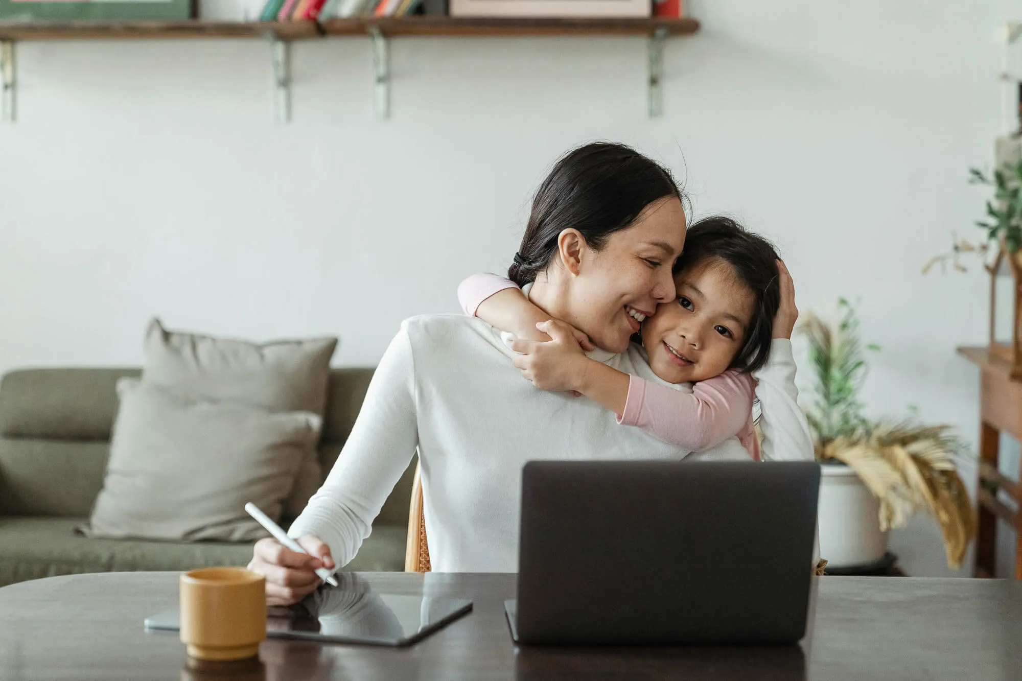 A woman works at a table with a laptop and stylus while a young girl hugs her from behind, both smiling.