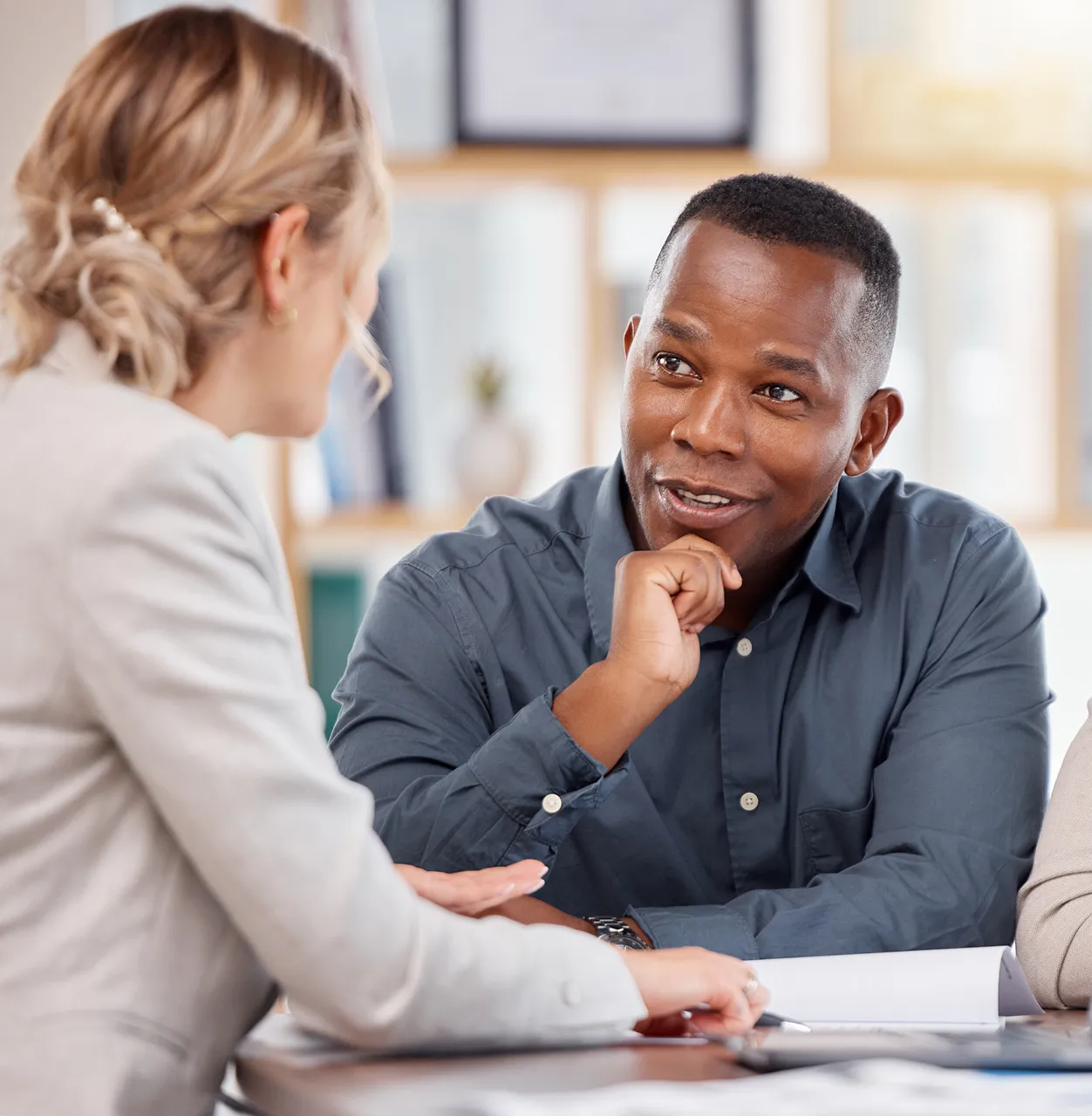 Two people sit at a table having a discussion, with documents and a pen in front of them in an office setting.