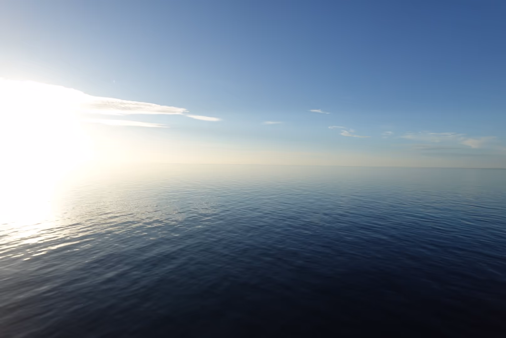 Calm ocean water under a clear blue sky with the sun shining near the horizon.
