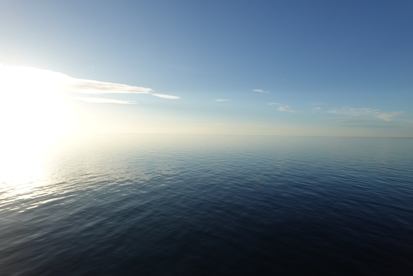 Calm ocean water under a clear blue sky with the sun shining near the horizon.