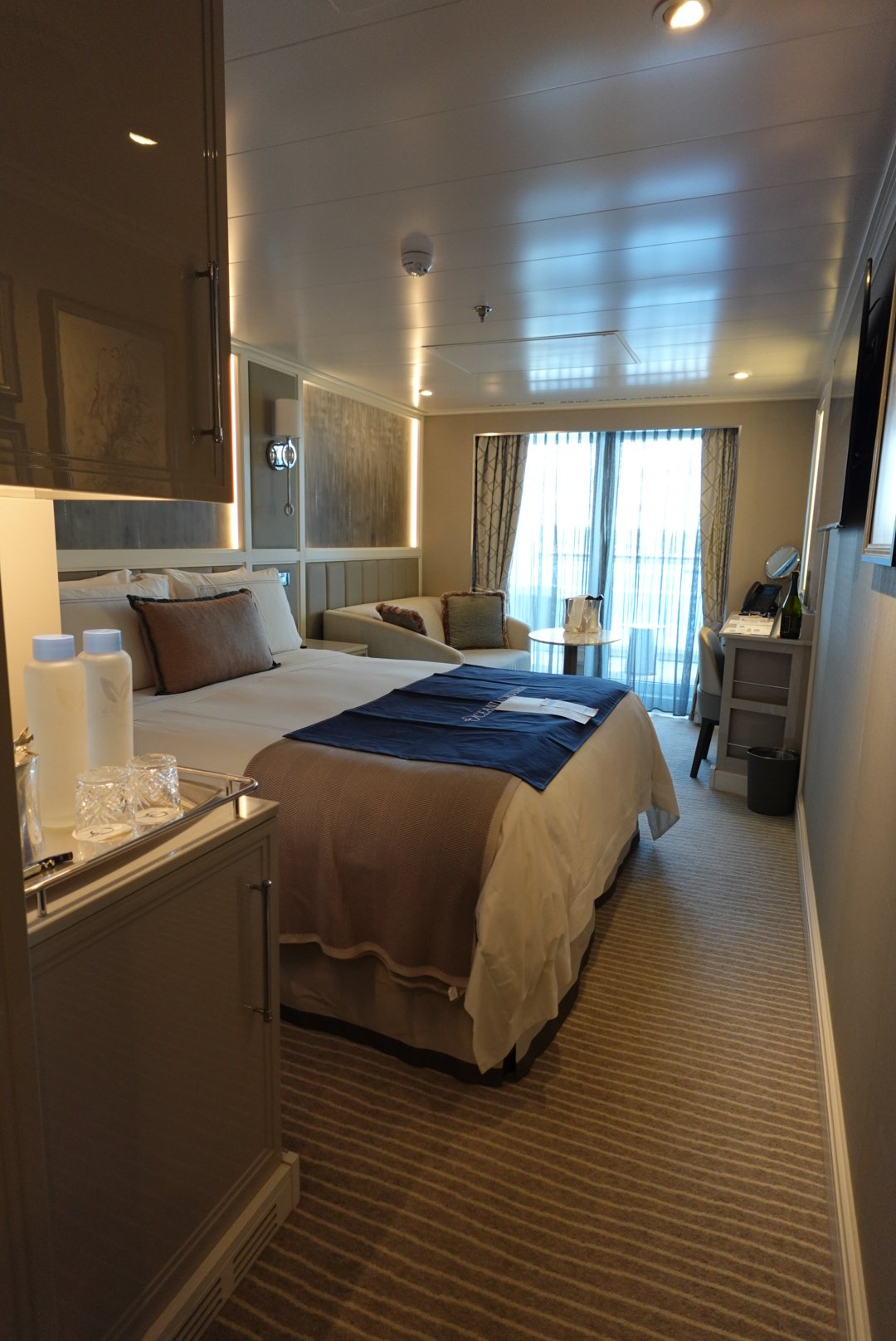 Modern hotel room with a bed, beige sofa, desk, and a large window with sheer curtains.