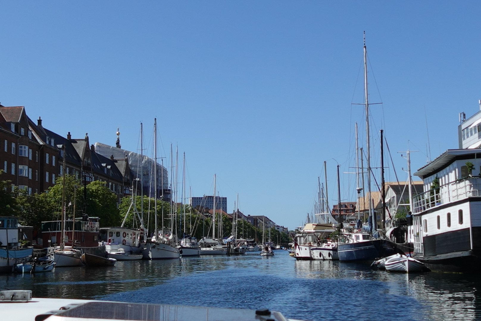 View down a canal lined with docked sailboats and buildings under a clear blue sky.