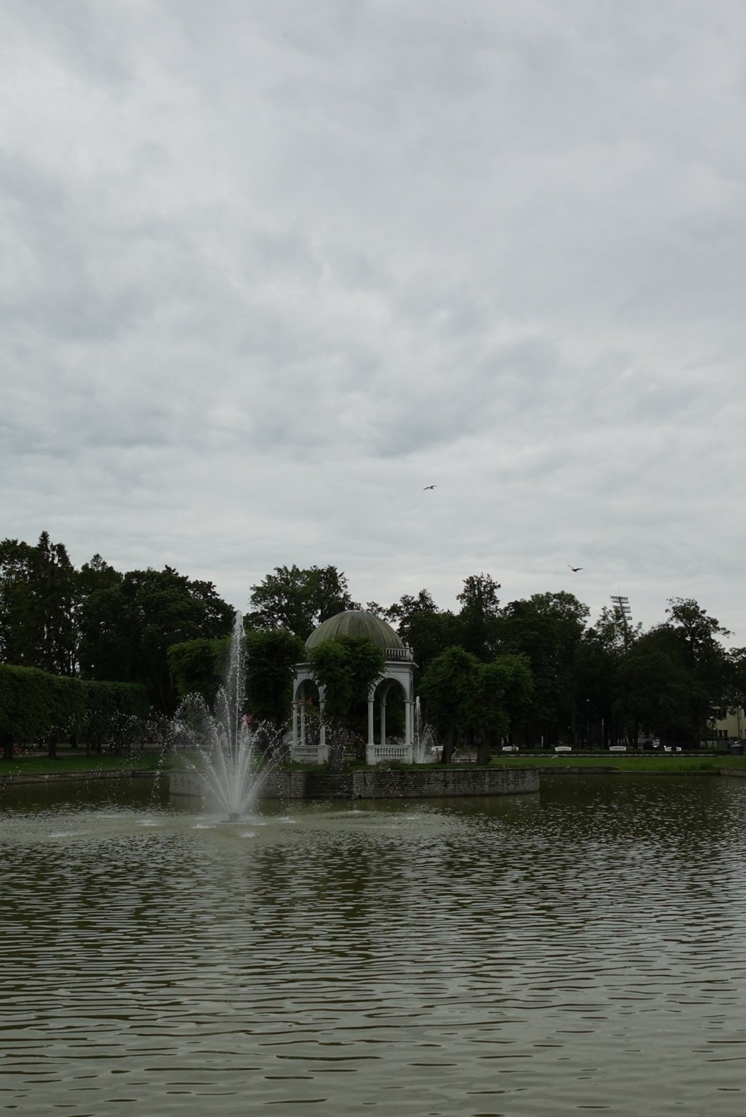 White gazebo with a green dome roof on a small island in a pond with a water fountain spraying, surrounded by trees under a cloudy sky.