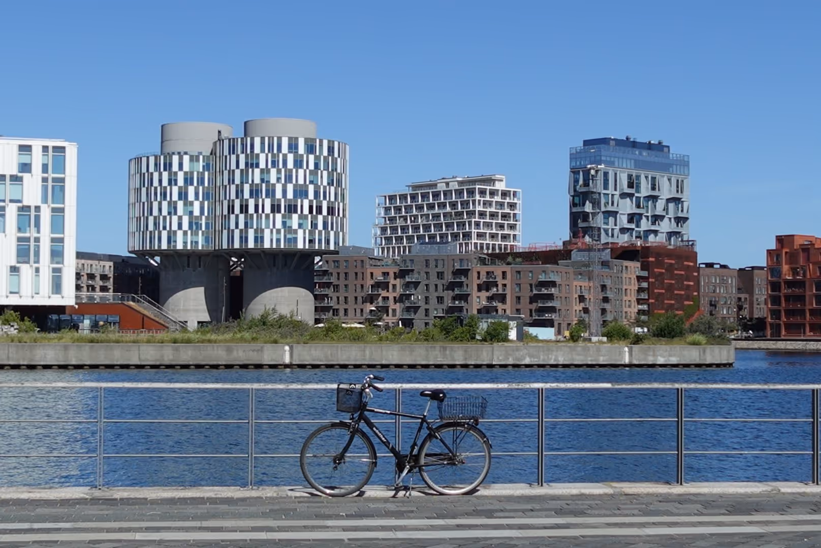 Black bicycle parked against a metal railing by a waterfront with modern multicolored buildings in the background under a clear blue sky.