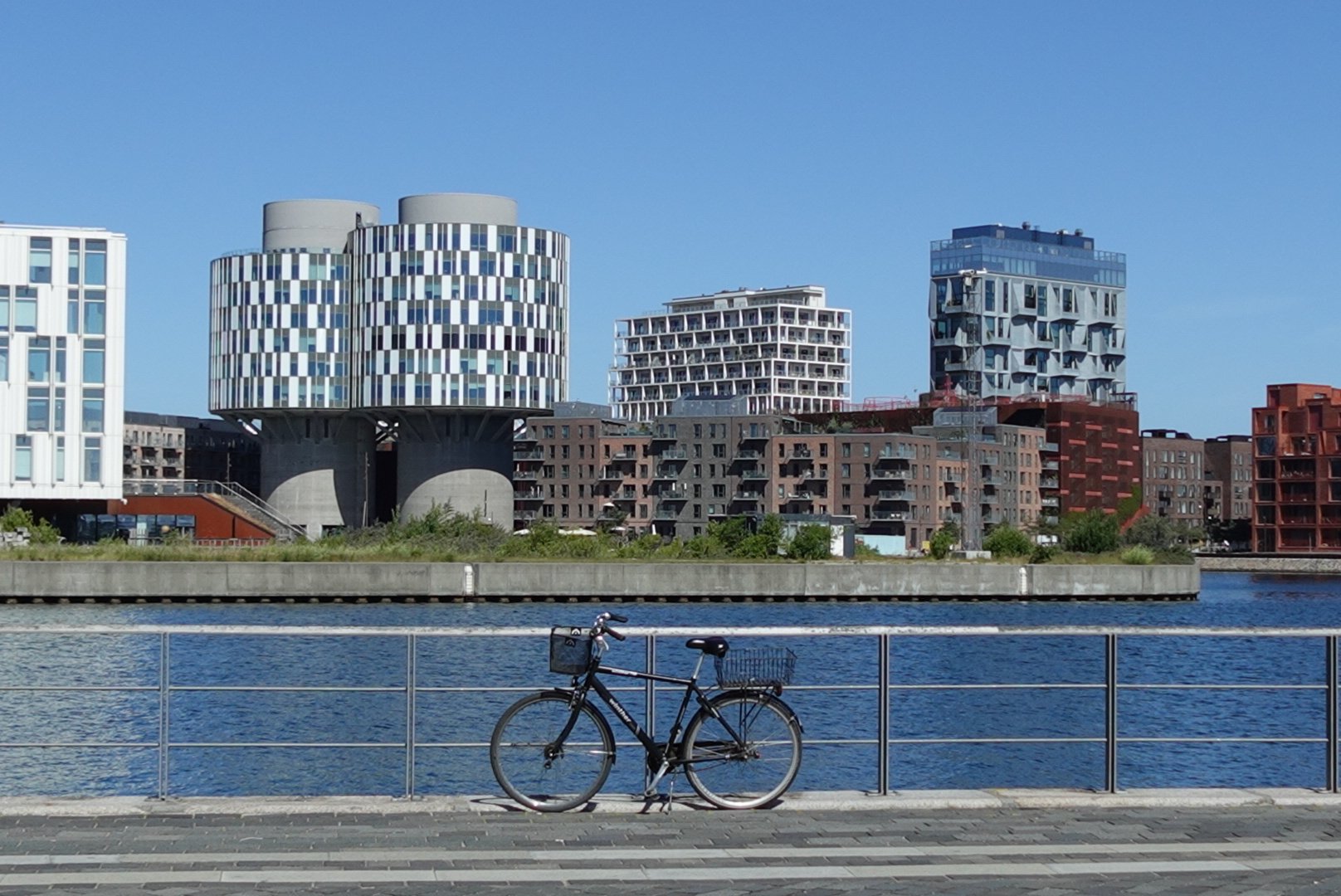 Black bicycle parked against a metal railing by a waterfront with modern multicolored buildings in the background under a clear blue sky.
