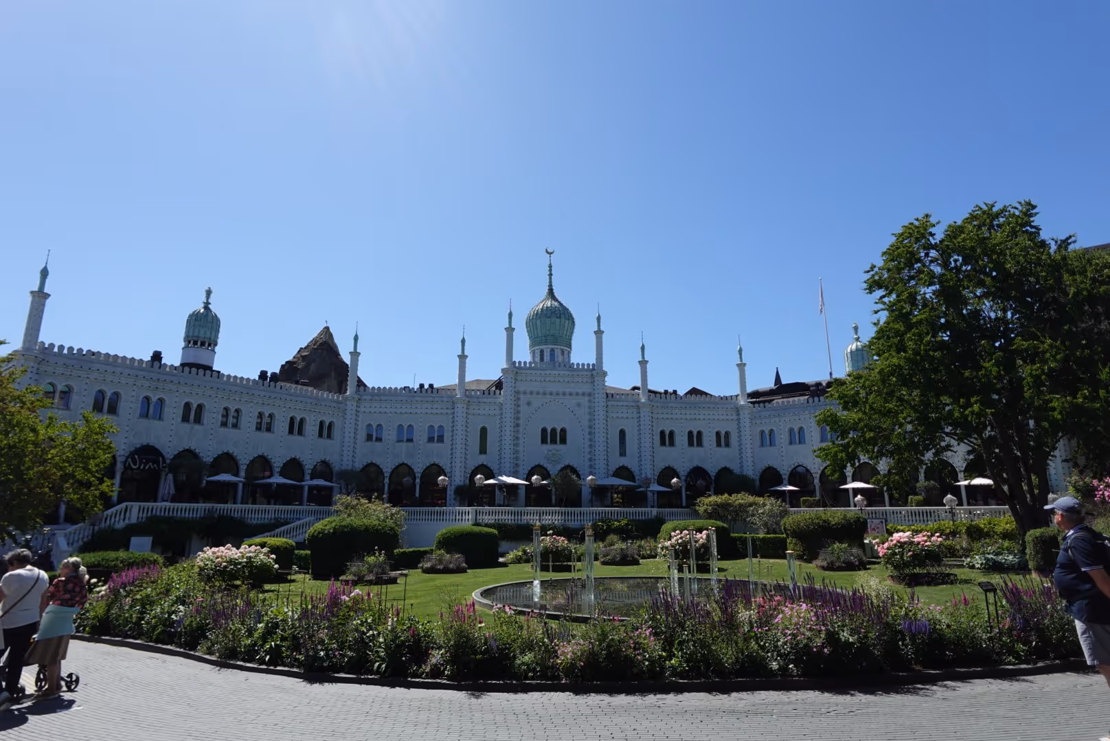Wide view of a white ornate building with green domes and minarets, surrounded by a garden with flowers and a fountain under a clear blue sky.