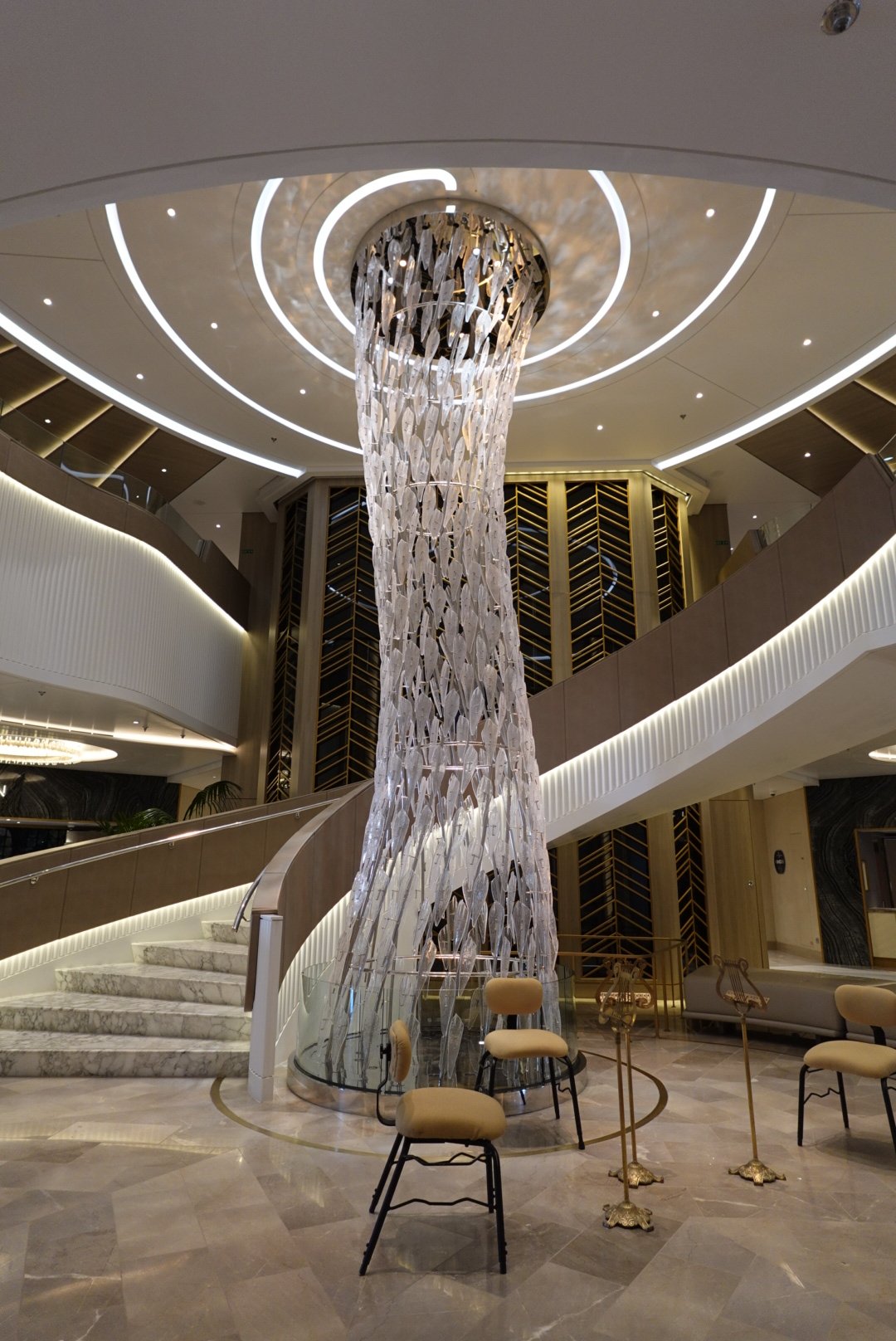 Modern interior with a tall, sculptural glass chandelier surrounded by beige chairs and marble stairs.