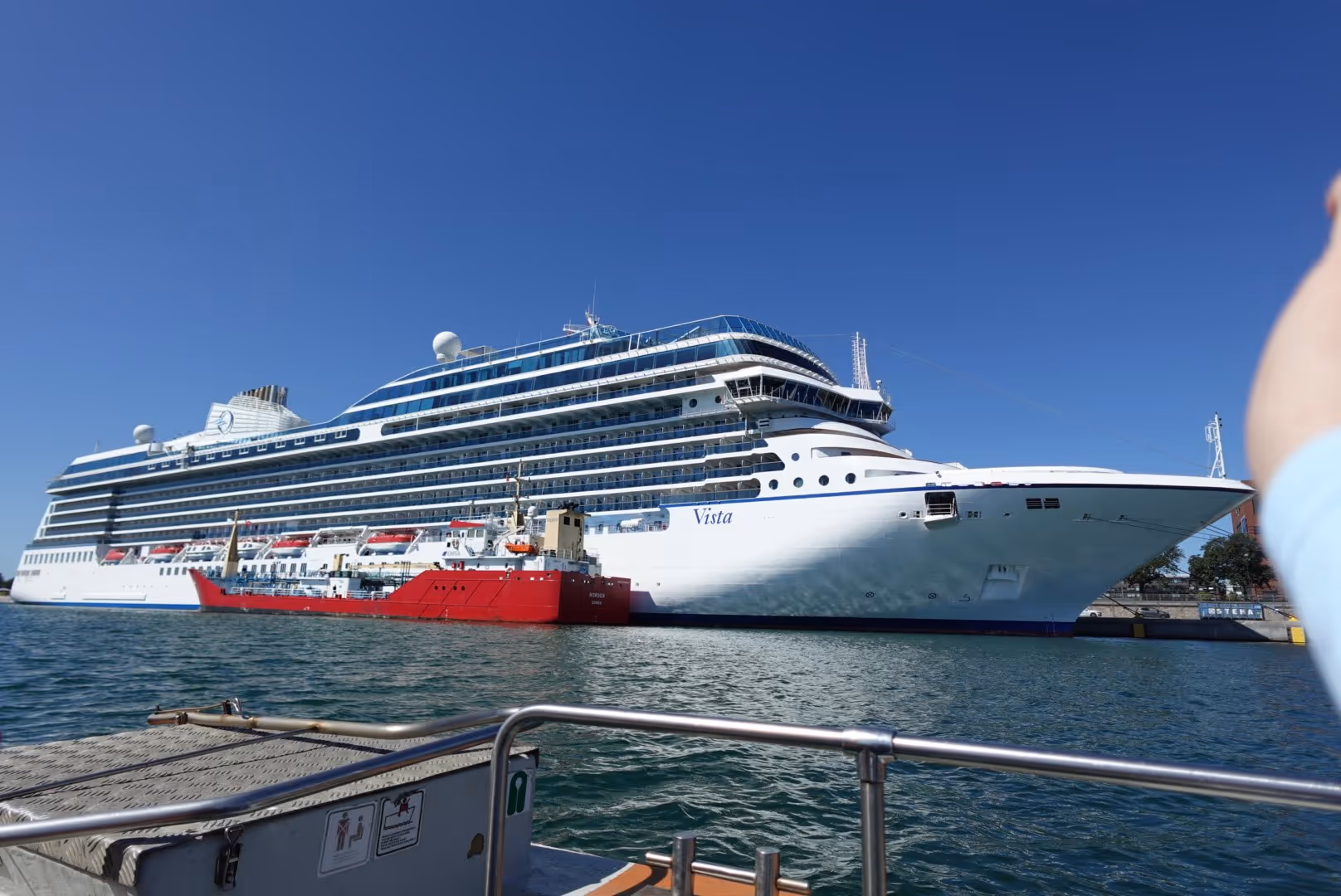 Large white cruise ship named Vista docked at a harbor with a red supply vessel alongside.